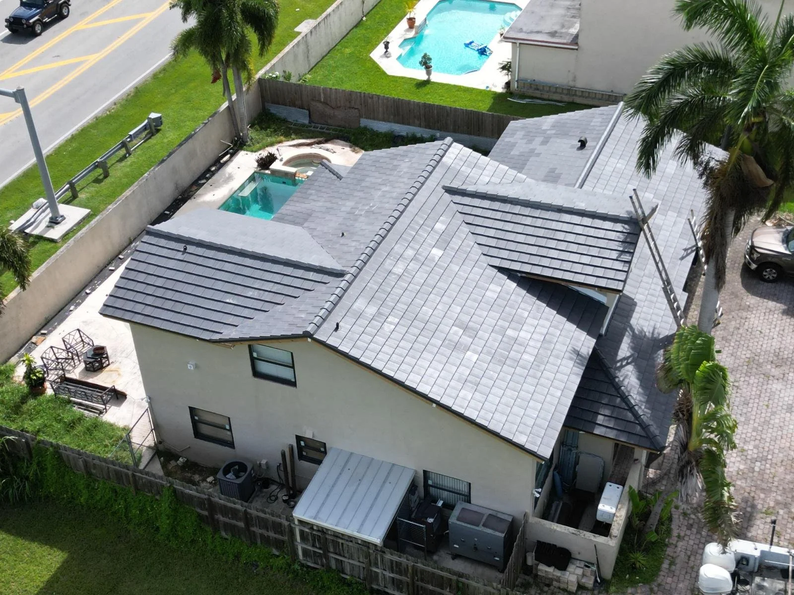 Aerial view of a house with a gray tiled roof, backyard with two swimming pools, a hot tub, palm trees, and outdoor seating, surrounded by a wooden fence.