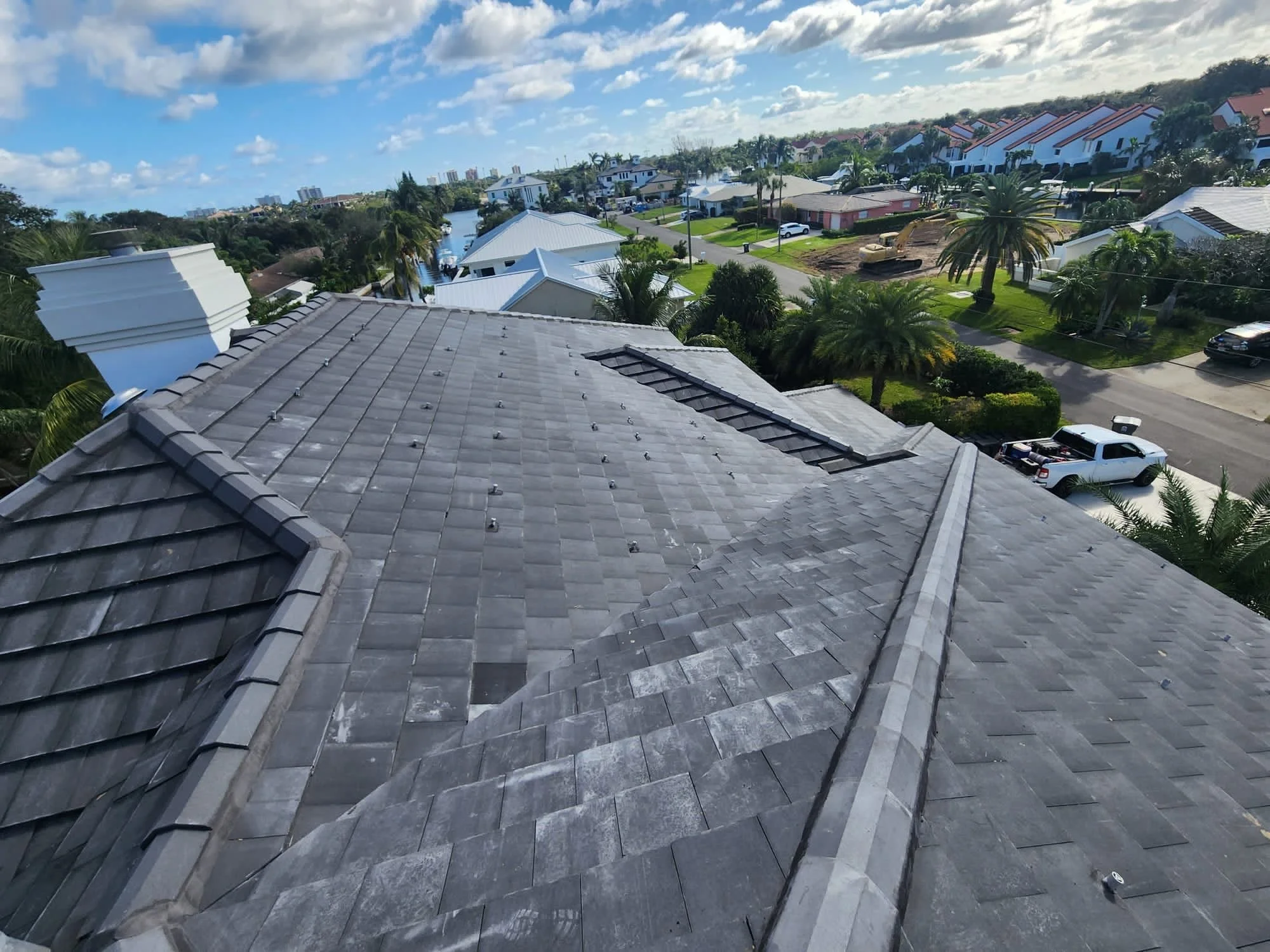 Aerial view of a residential neighborhood showing gray tiled rooftops, palm trees, and streets with parked cars under a partly cloudy sky.