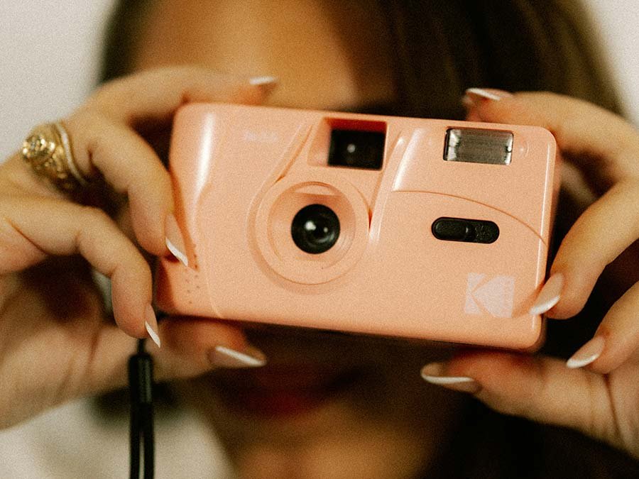 A person holding a peach-colored instant camera in front of their face, with their hands on either side of the camera. The person's nails are painted white, and they are wearing rings on their fingers.