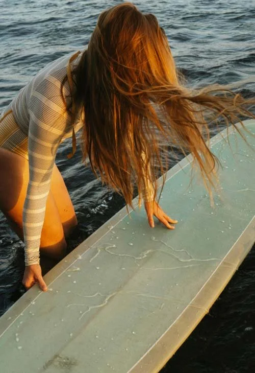 A person with long, flowing hair leaning on a surfboard in the water, preparing to surf.