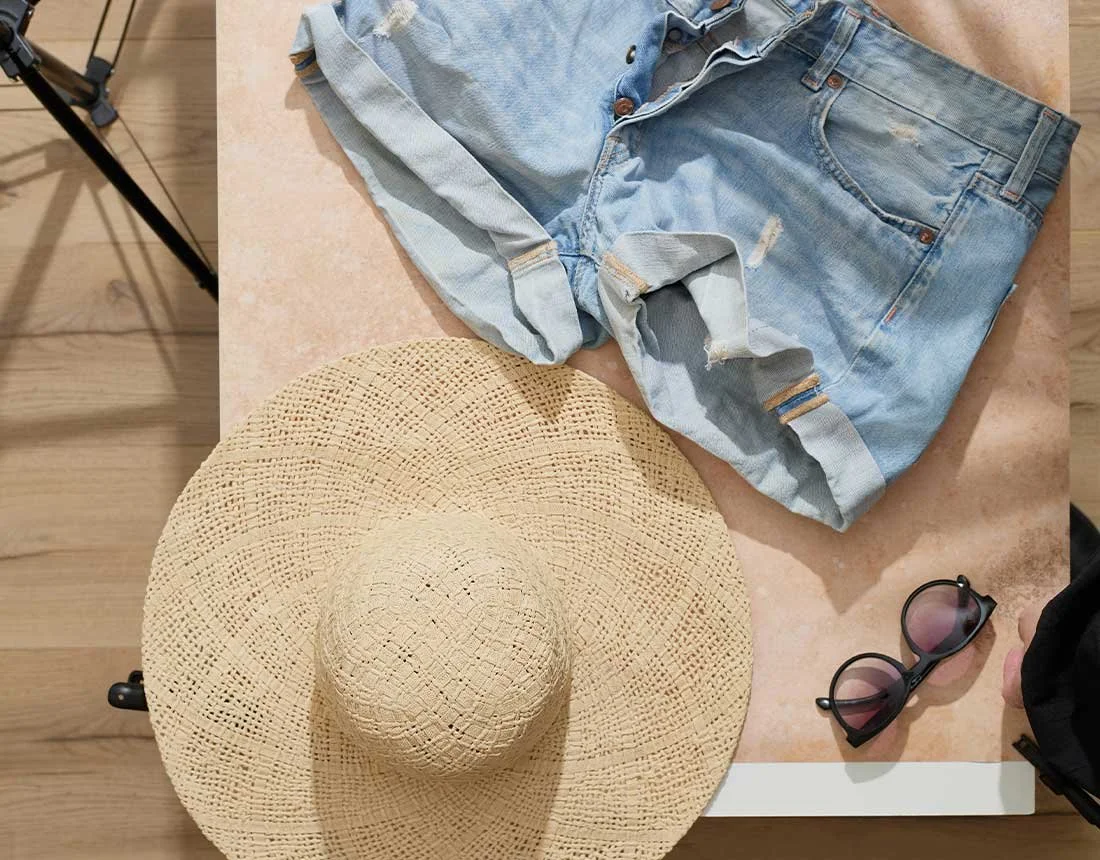 A pair of ripped denim shorts, a large woven sun hat, sunglasses, and a black backpack on a table.