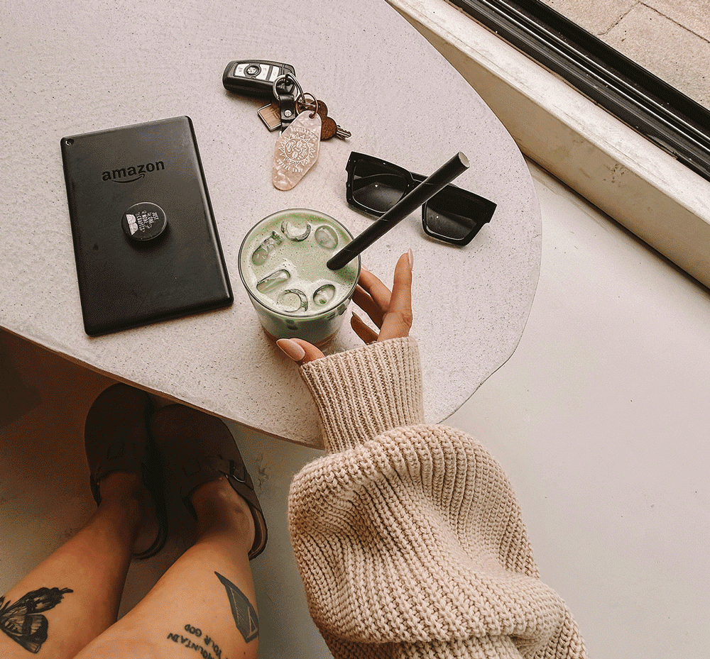 Overhead view of a table with a hand holding a glass of iced matcha latte, a smartphone, car keys, sunglasses, and a Kindle e-reader. A person wearing a beige chunky knit sweater and black boots is sitting nearby.