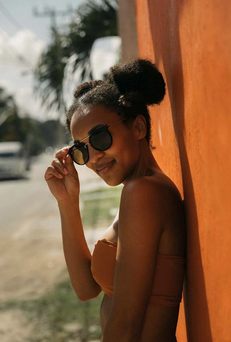 A woman leaning against an orange wall outdoors, wearing dark sunglasses and an orange strapless top, smiling and adjusting her sunglasses.