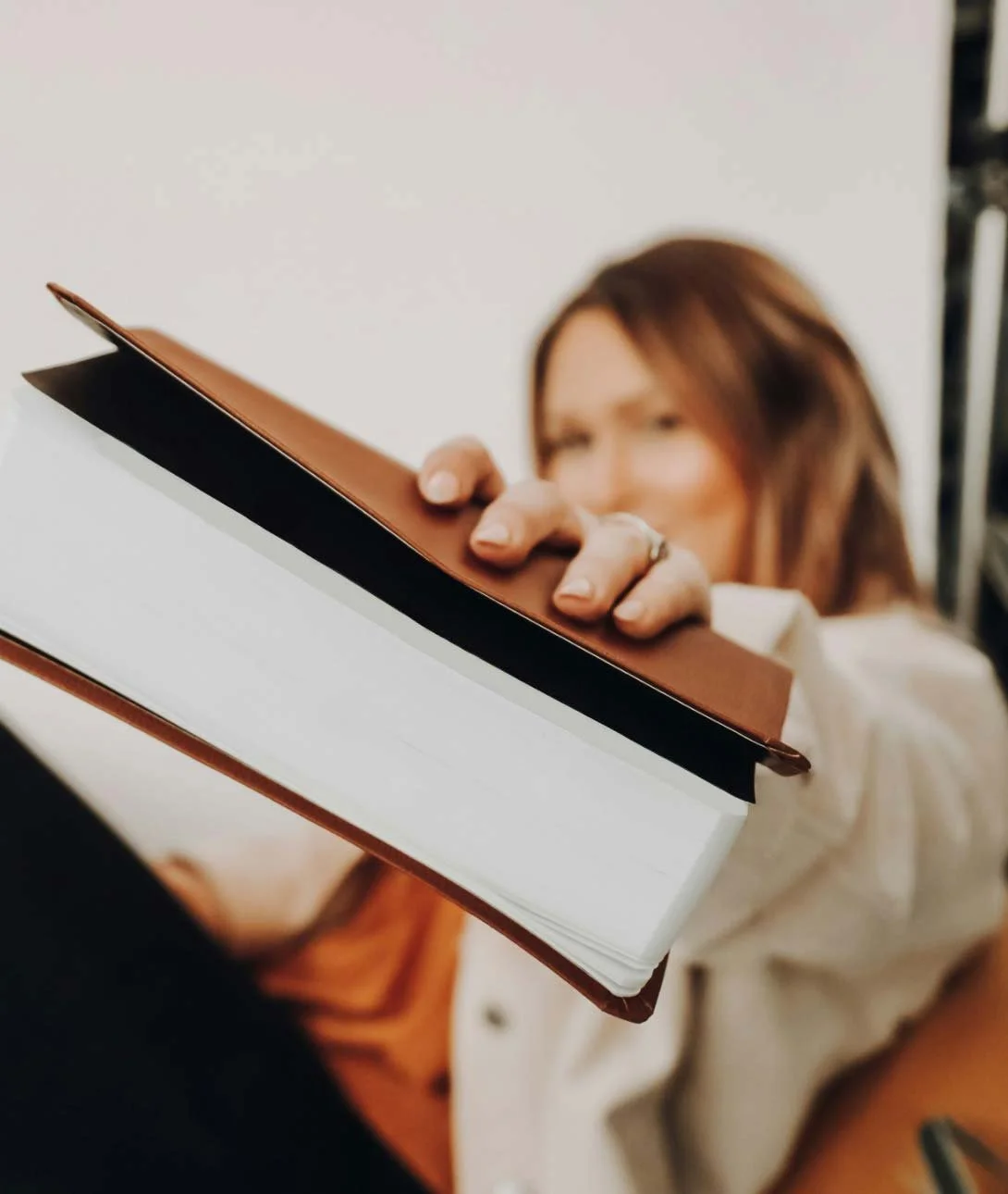 Person holding an open leather-bound notebook or planner, with their face partially visible and out of focus in the background.
