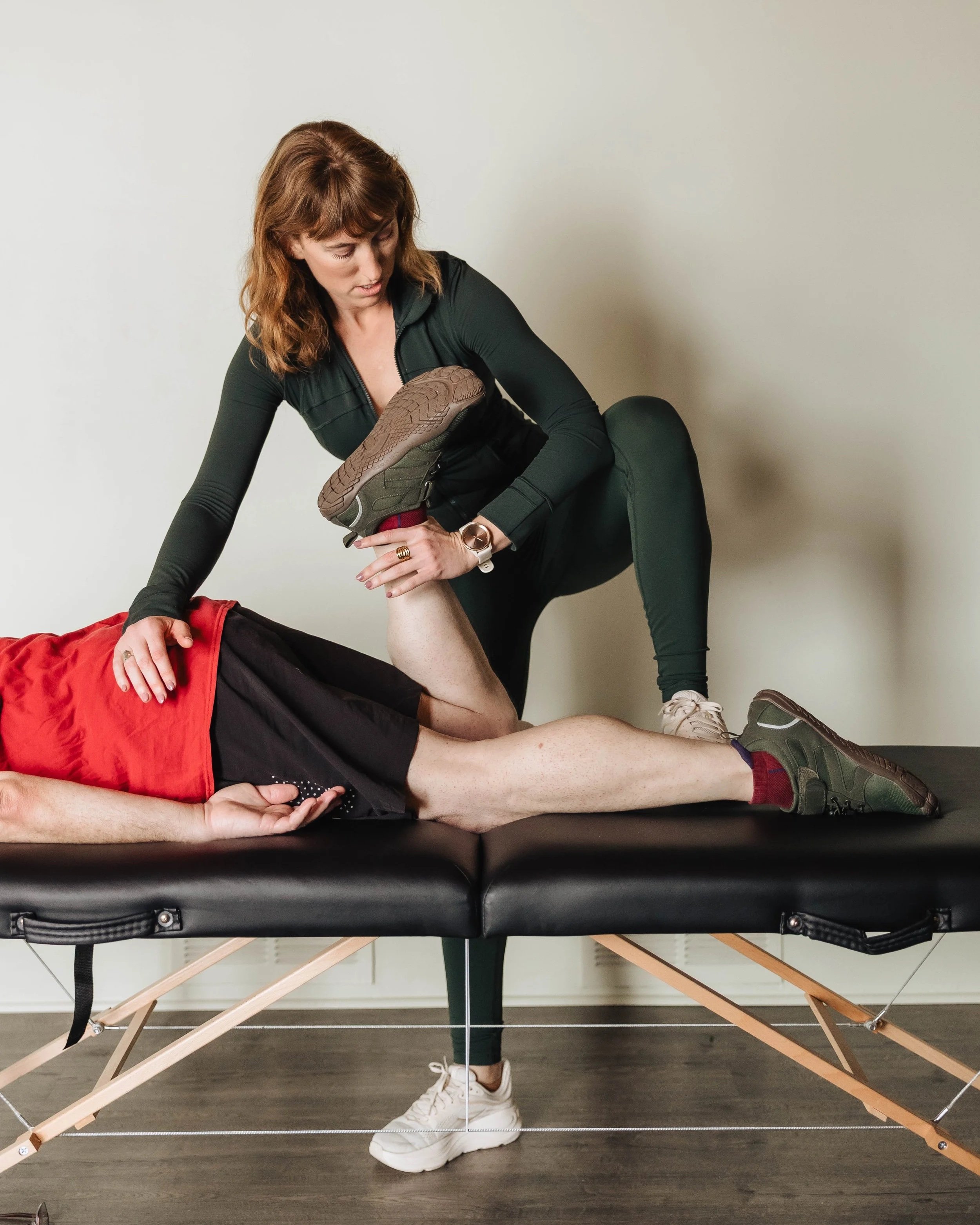 A woman demonstrating a stretch on a massage table with another person lying underneath, holding their knee while the woman pulls the other leg, in a clinical setting.