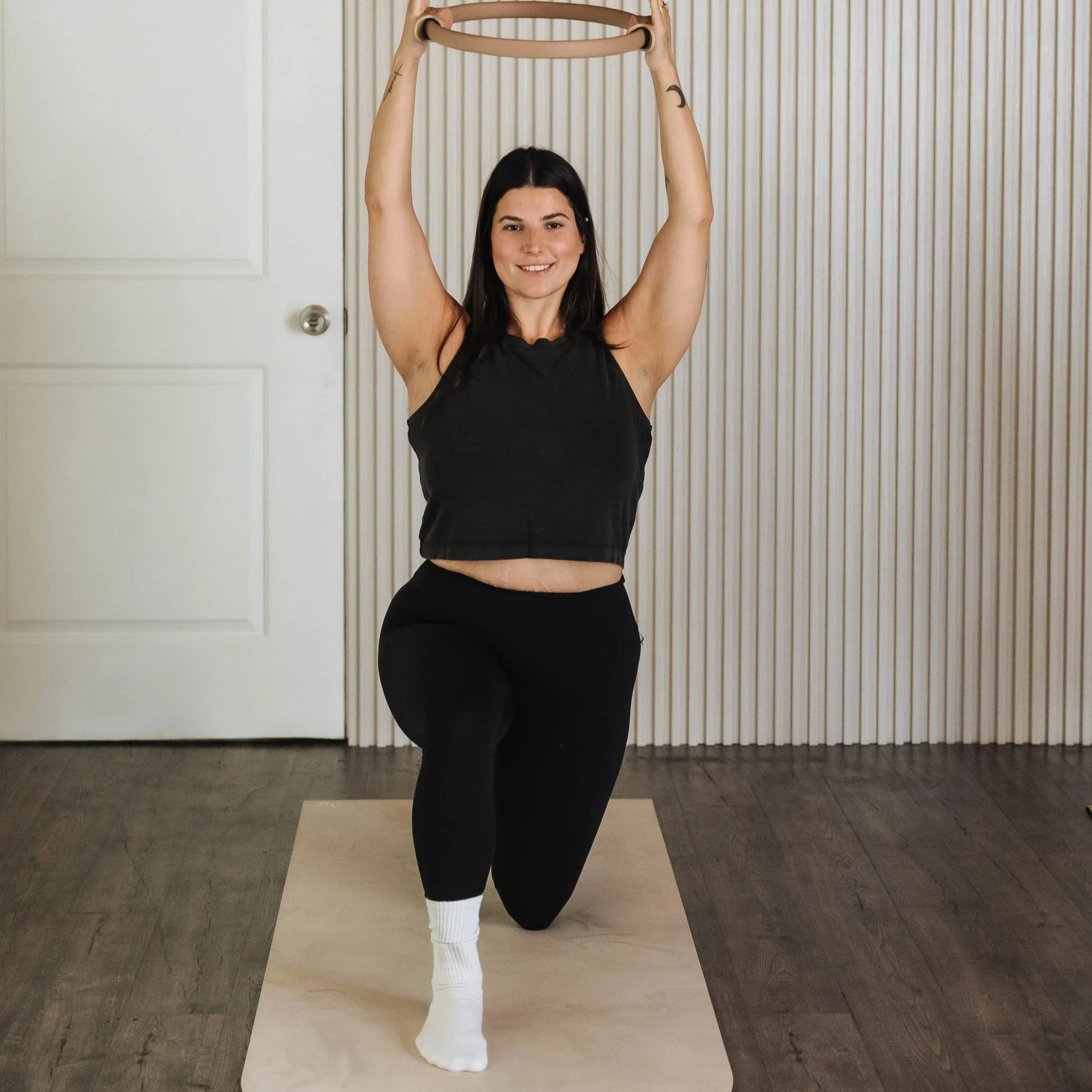 A woman with long dark hair, wearing a black sleeveless top and black pants, is practicing yoga indoors against a background of a closed door and vertical striped wall. She is in a balancing pose, standing on one leg with the other knee bent and foot resting on her standing thigh. She holds a wooden yoga ring with both hands raised overhead.