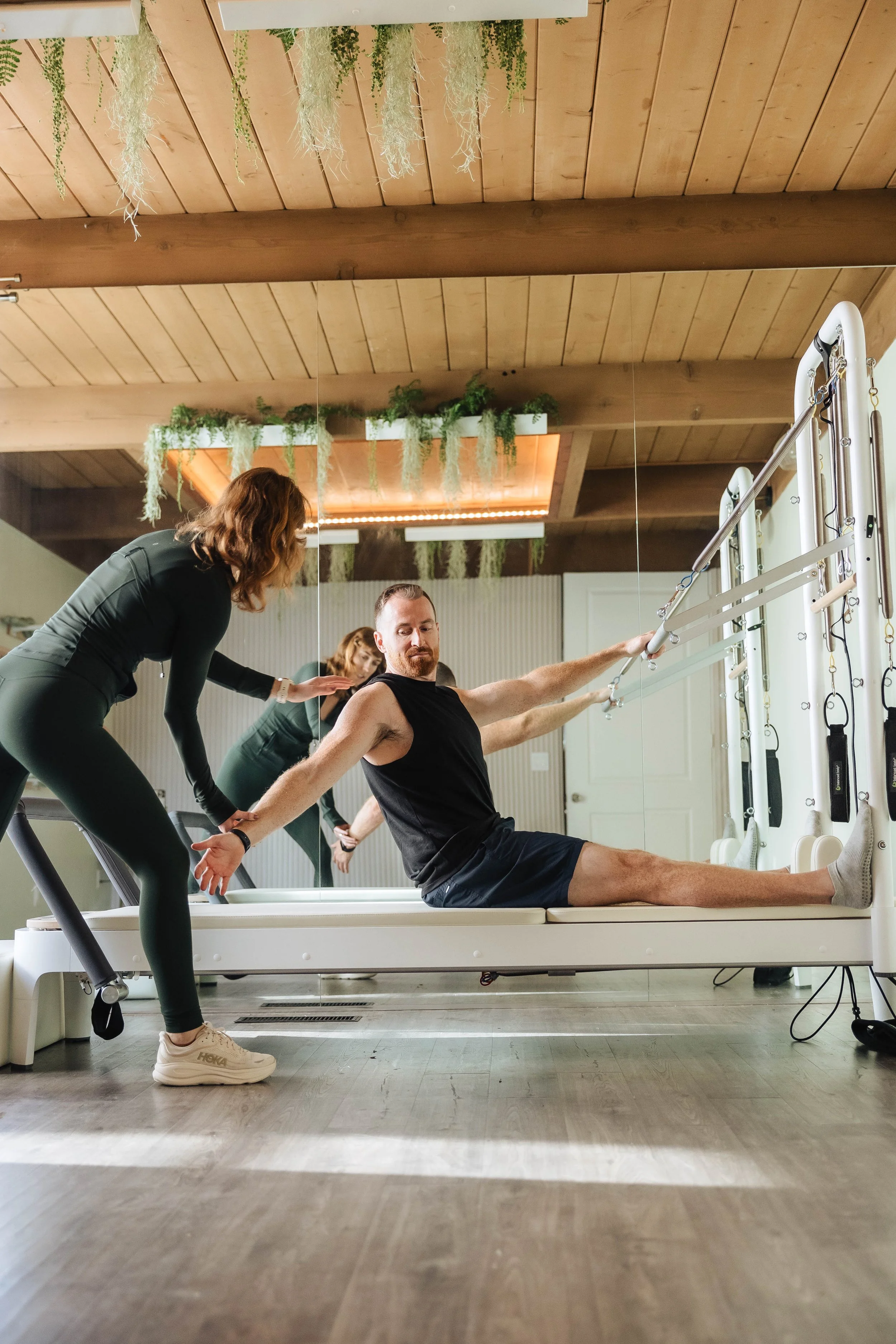 A physical therapist assisting a man during Pilates exercises in a gym with wooden ceiling and plants hanging from the ceiling.