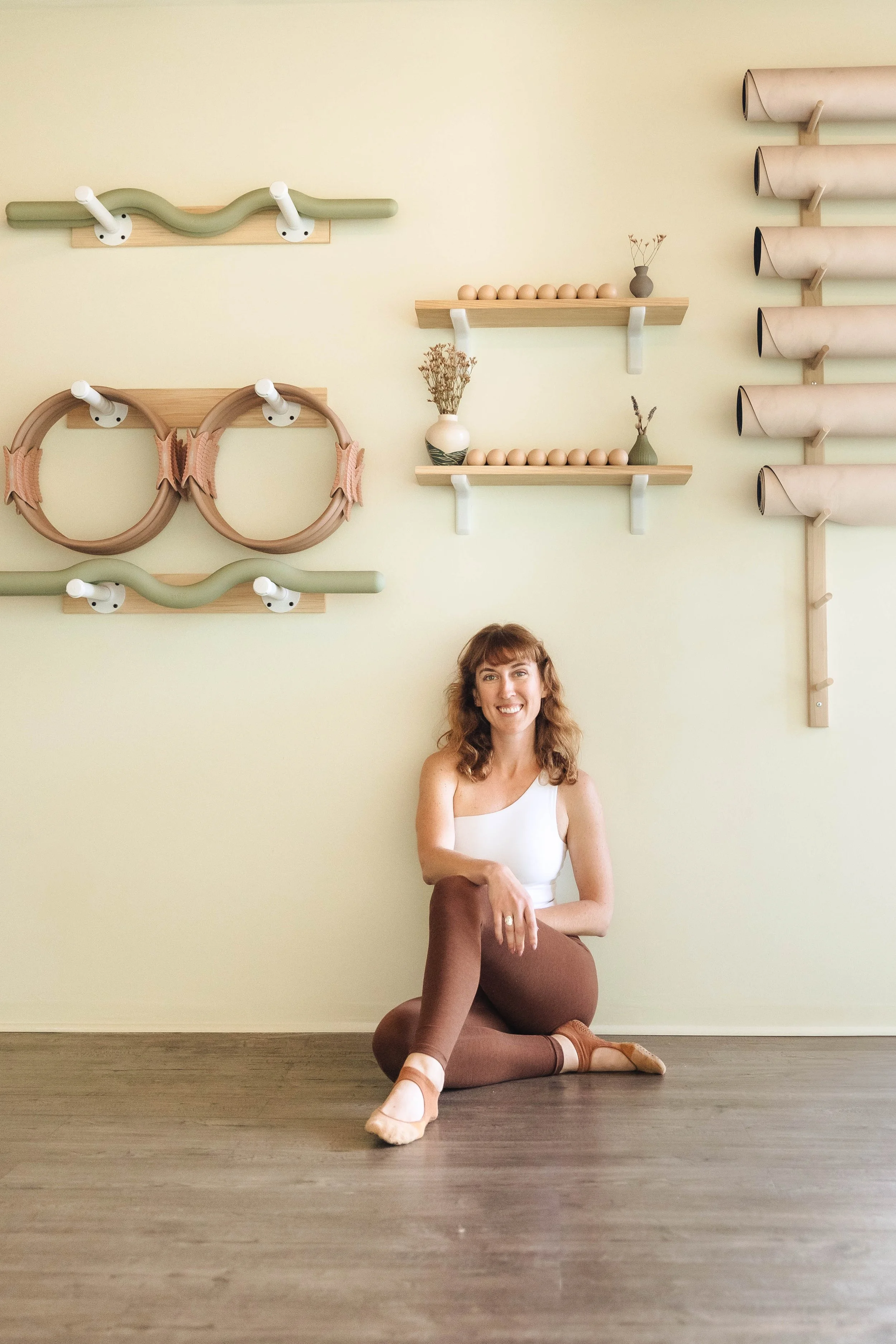 A woman with curly hair smiling, sitting on the floor with one arm resting on her knee, in a room with light yellow walls and wooden flooring. Behind her, there are minimalist wall-mounted shelves with decorative items including vases, dried flowers, and wooden balls, along with some wall hooks and cylindrical wall-mounted storage.