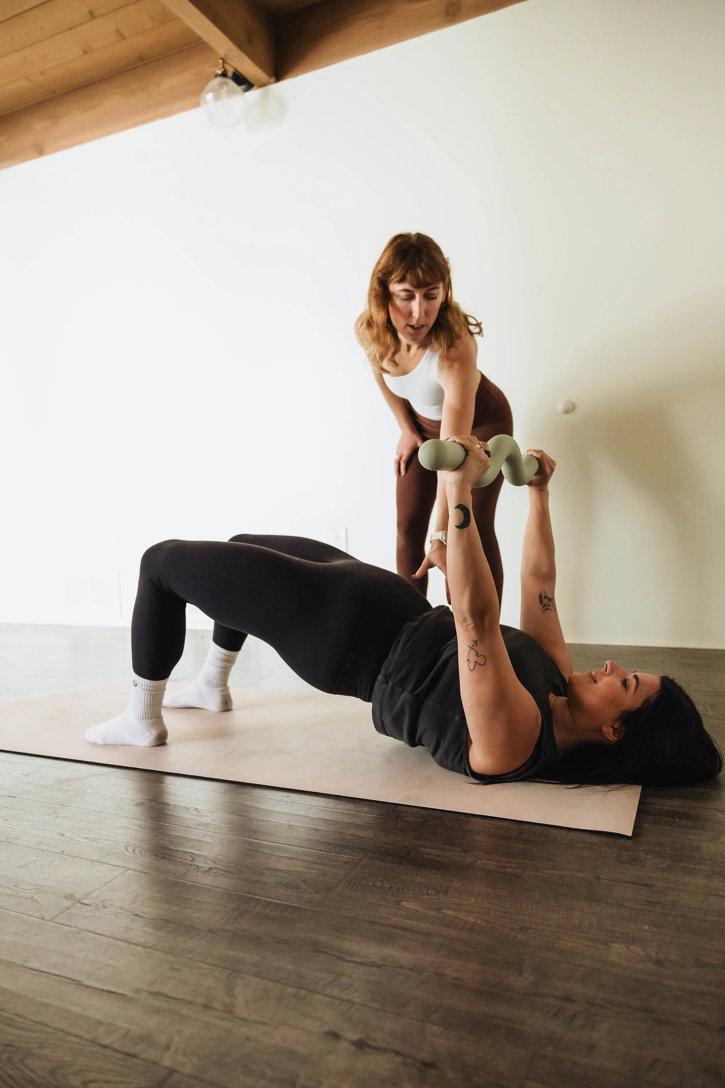 Two women are practicing yoga in a bright room. One woman is lying on her back holding a pair of dumbbells, while the other woman stands beside her, offering assistance. The scene reflects a workout or yoga session using a mirror to capture their activity.