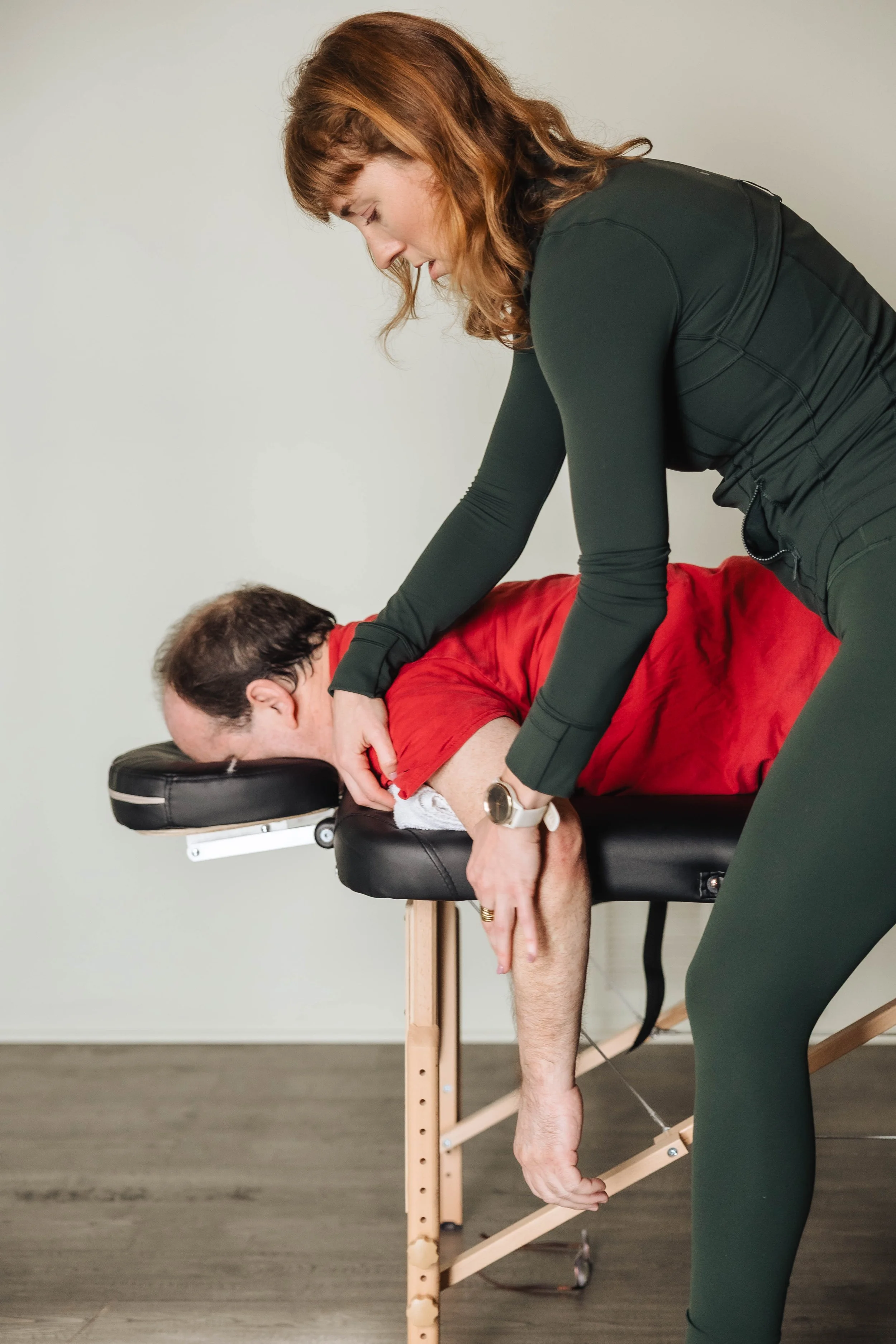 A woman practicing yoga or stretching with a man in a red shirt lying face down on a massage table, in a room with light-colored walls.