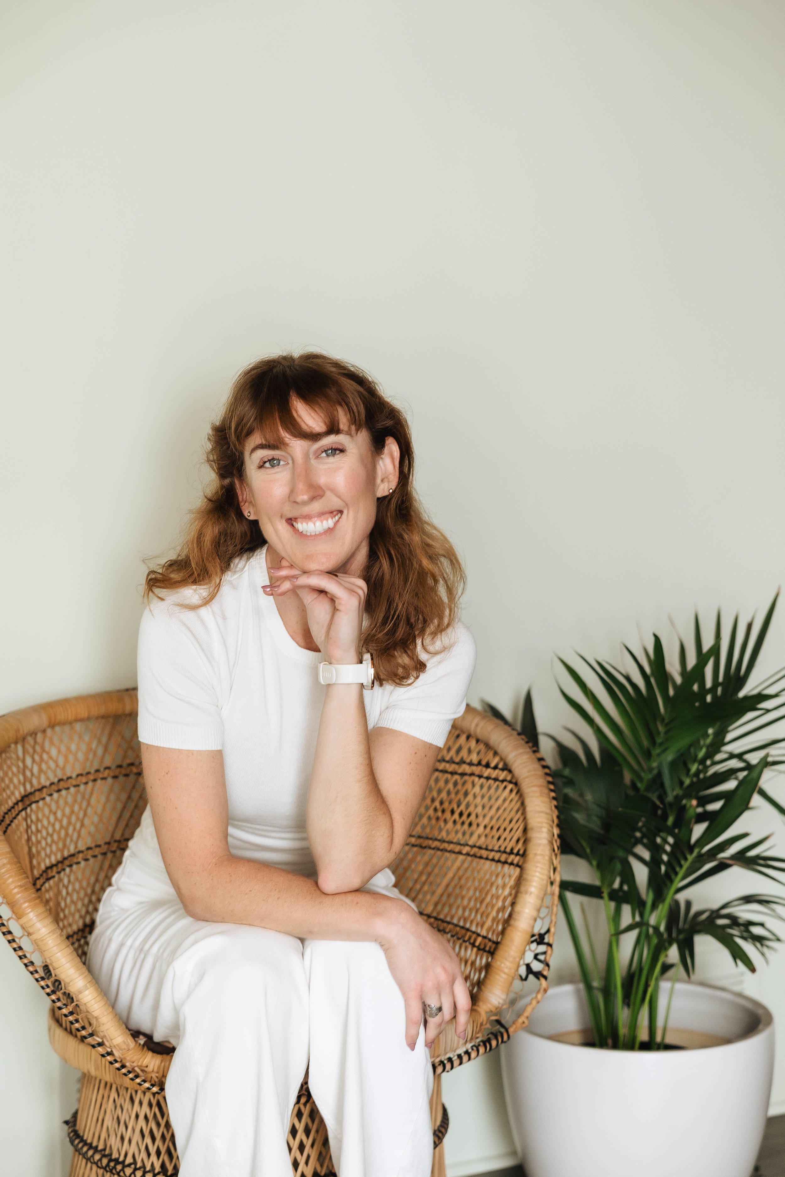A smiling woman with curly red hair and light skin sitting on a wicker chair, wearing a white short-sleeved top and white pants, with her hand resting under her chin. There is a large green potted plant next to her.