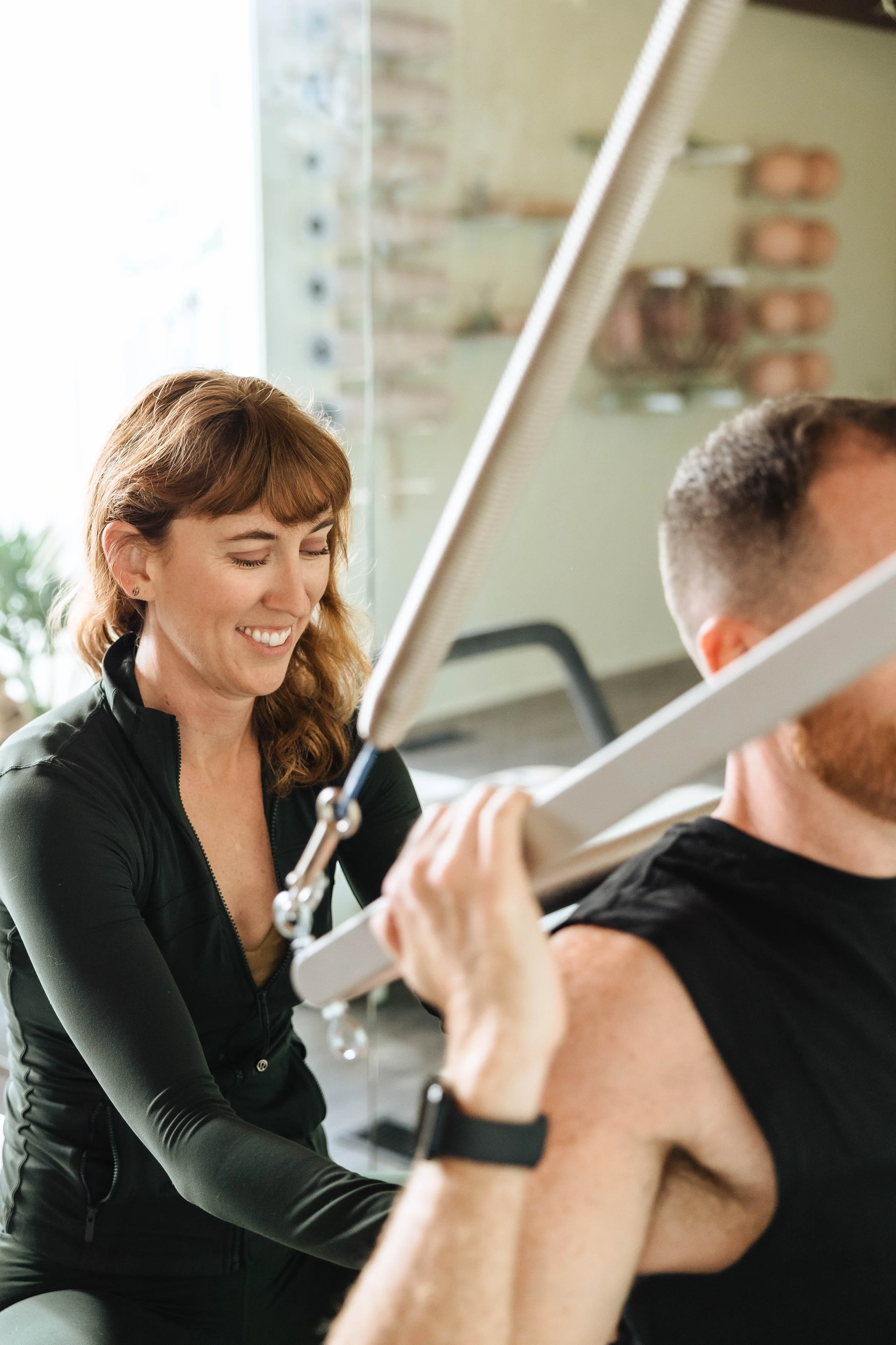 A woman with red hair smiling while assisting a man with an arm workout in a gym, with weights visible in the background.