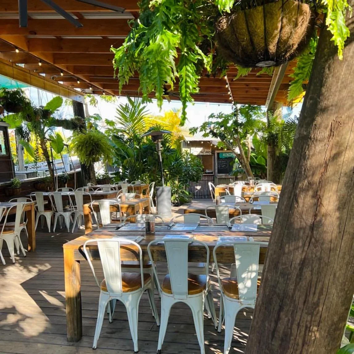 Kokomo Kitchen and Bar Gold Coast. Outdoor restaurant patio with wooden tables and white metal chairs, surrounded by lush green plants and hanging ferns under a wooden roof, with sunlight streaming in.