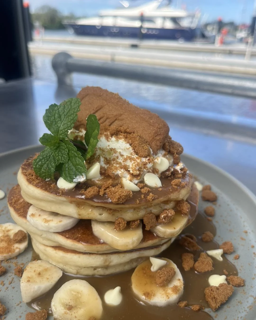 Kokomo Kitchen and Bar Gold Coast. Stack of pancakes topped with sliced bananas, whipped cream, caramel sauce, crumbled cookies, a cinnamon roll, and a sprig of mint, on a plate with a blurred marina in the background.