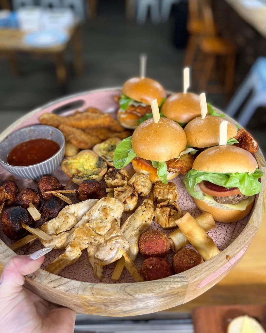 Kokomo Kitchen and Bar Gold Coast. A round serving tray with sliders, fried chicken wings, grilled chicken skewers, meatballs, fried spring rolls, and a bowl of dipping sauce, held by a hand.