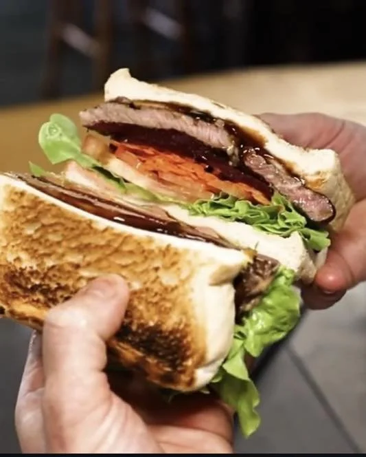 Kokomo Kitchen and Bar Gold Coast. Close-up of a person holding a layered sandwich with lettuce, tomato, beef patty, turkey, and melted cheese on toasted bread.