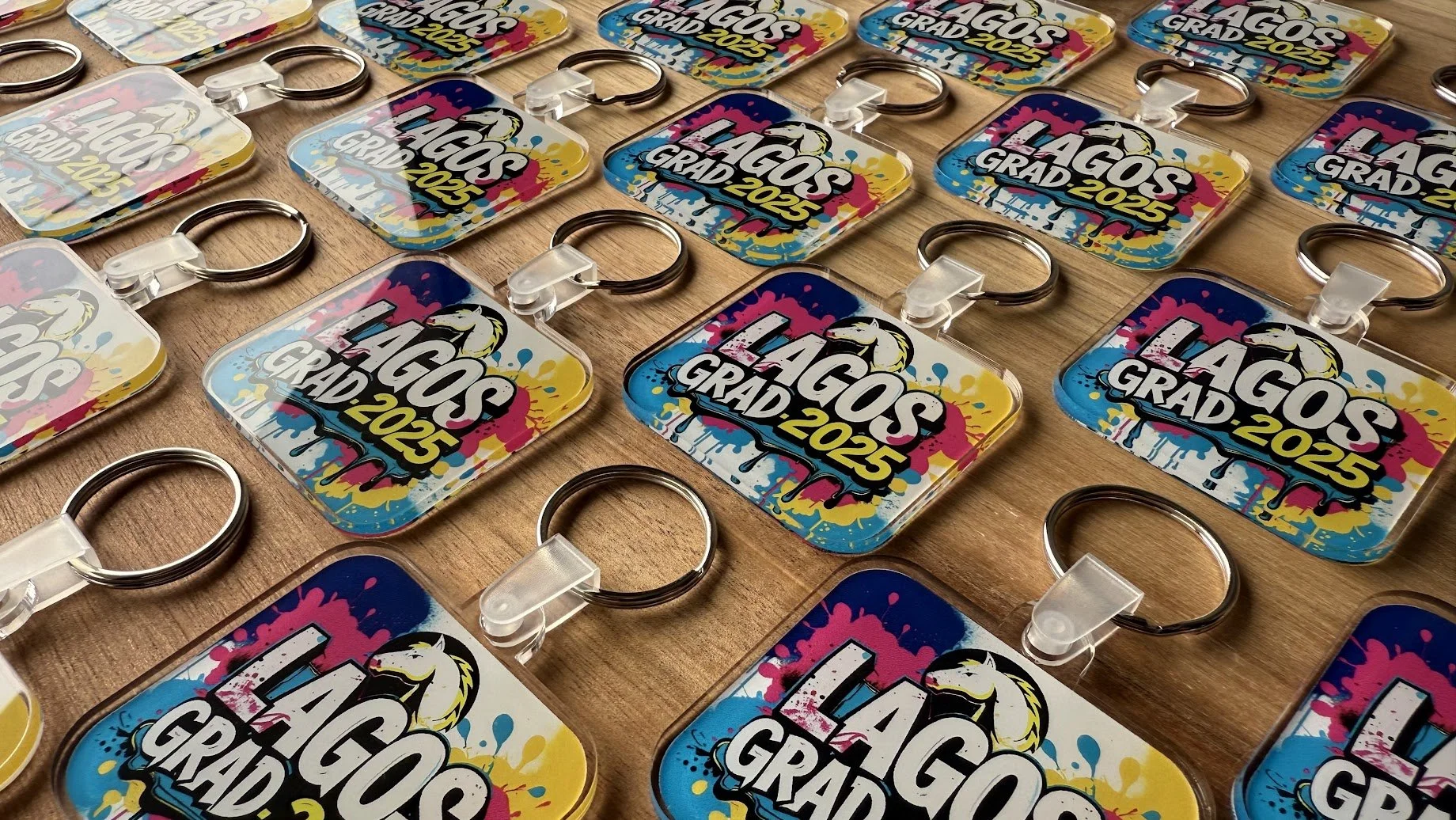 Custom Colorful keychains with a graffiti-style design and the words "Lagos Grad 2025" printed on them, spread out on a wooden surface.