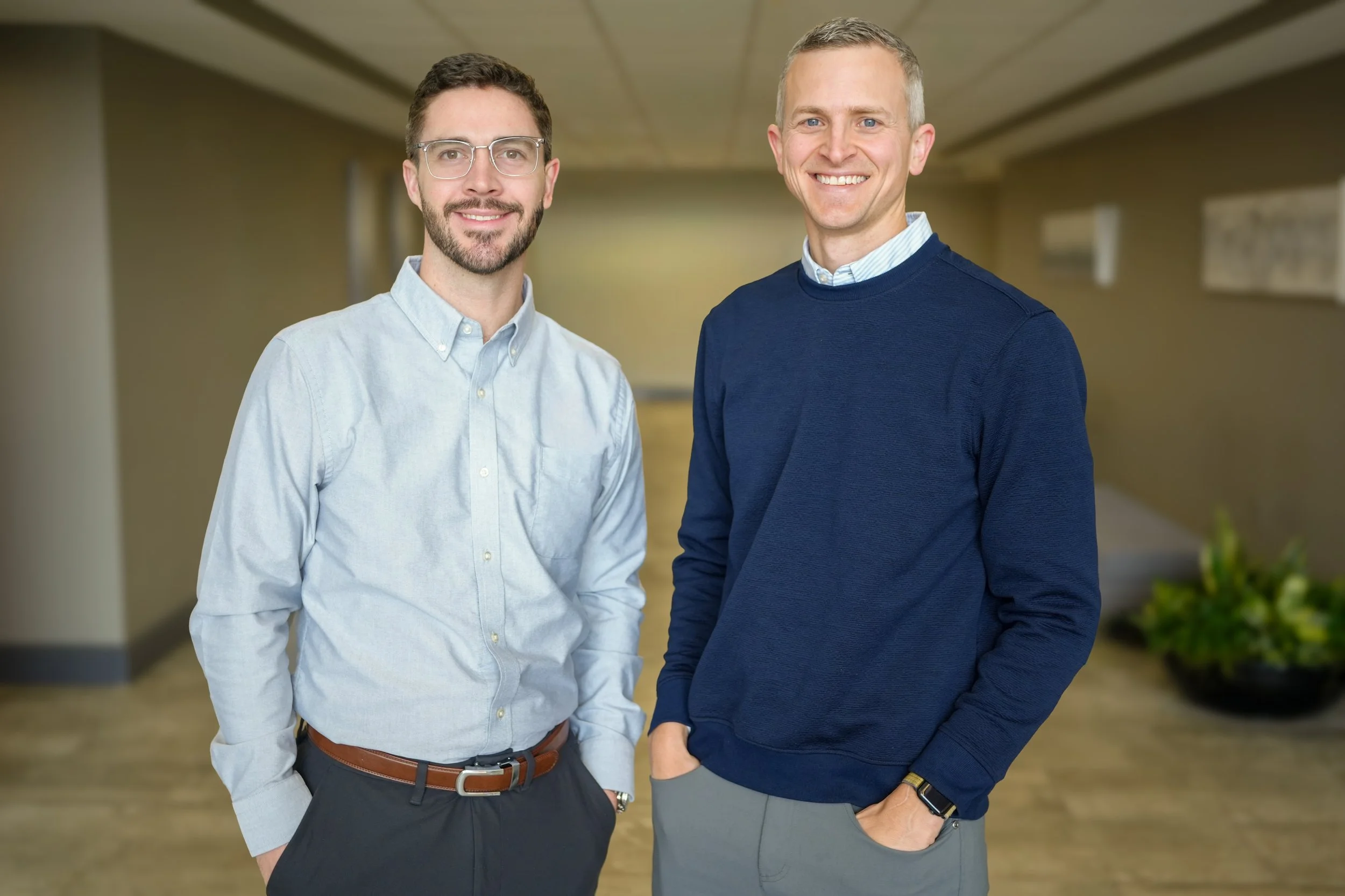 Two smiling men standing in an office hallway, one with glasses and a beard wearing a light blue shirt, and the other with short gray hair wearing a navy blue sweater and gray pants.