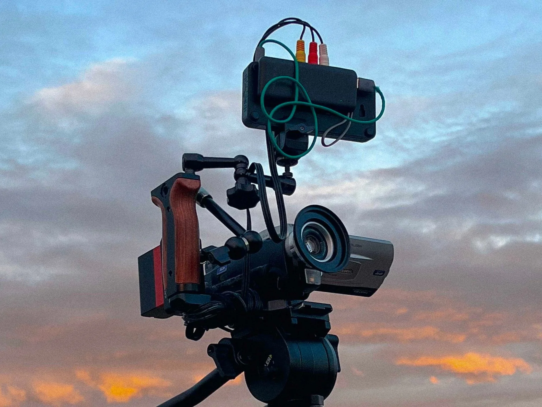A camera mounted on a tripod with additional electronic equipment attached, against a sunset sky with clouds.