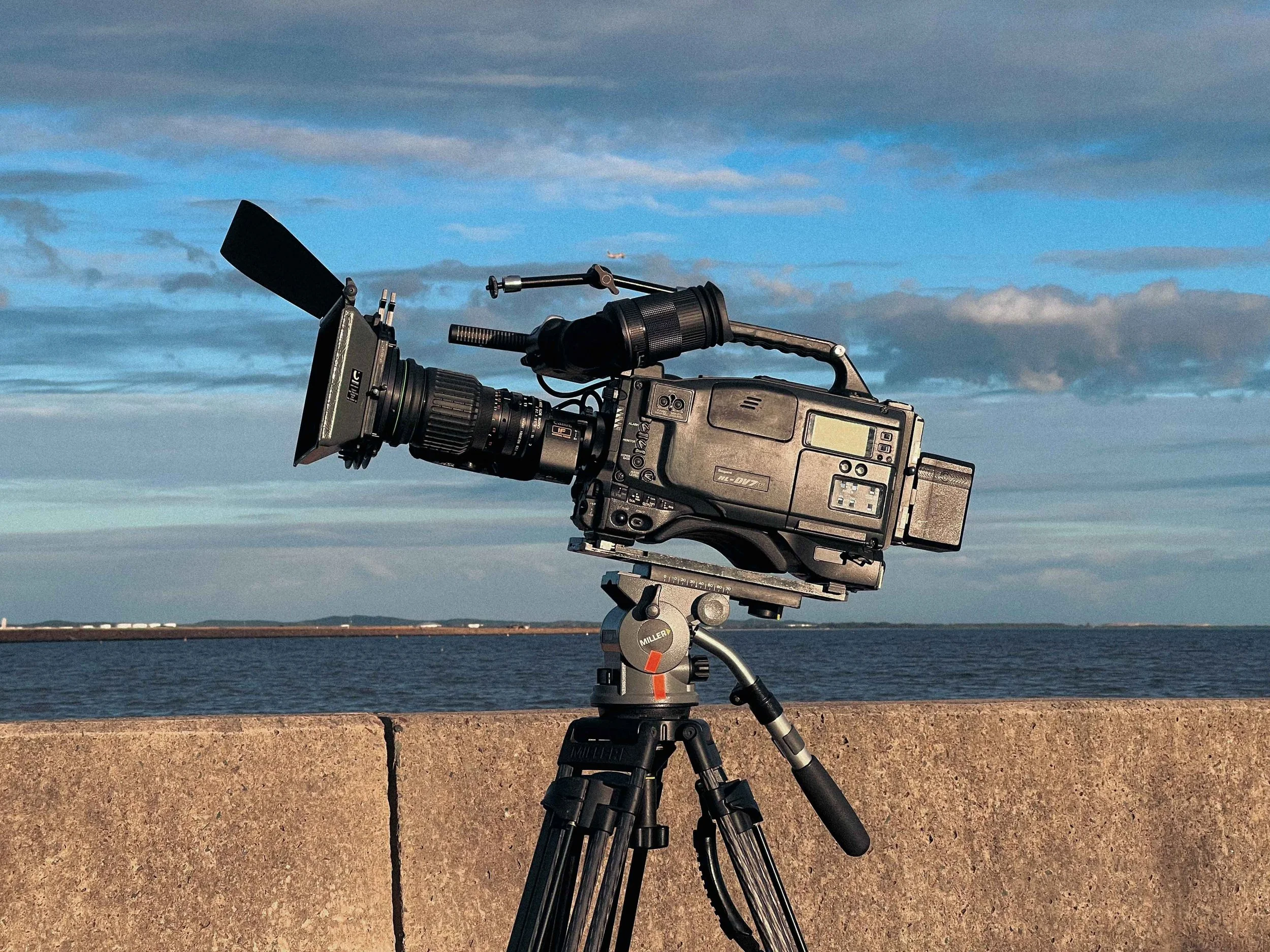 Professional video camera mounted on a tripod, positioned outdoors with a body of water and a cloudy sky in the background.