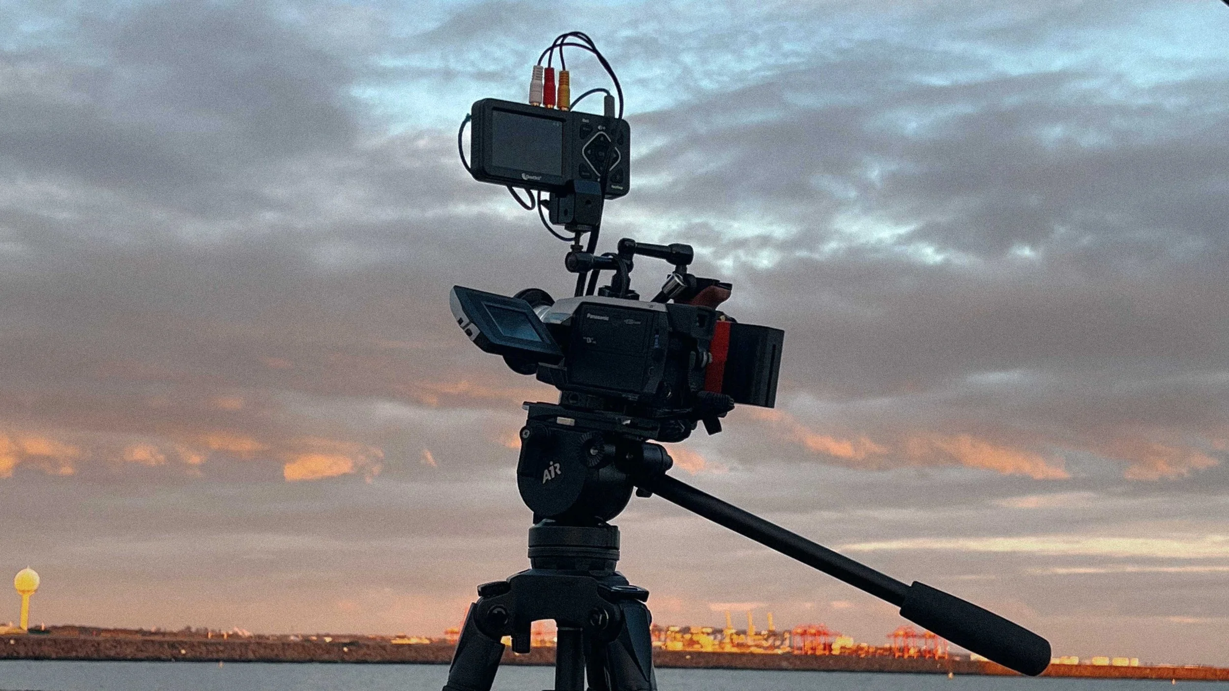 Camera mounted on a tripod facing a body of water with a city skyline and cloudy sky in the background at sunset.