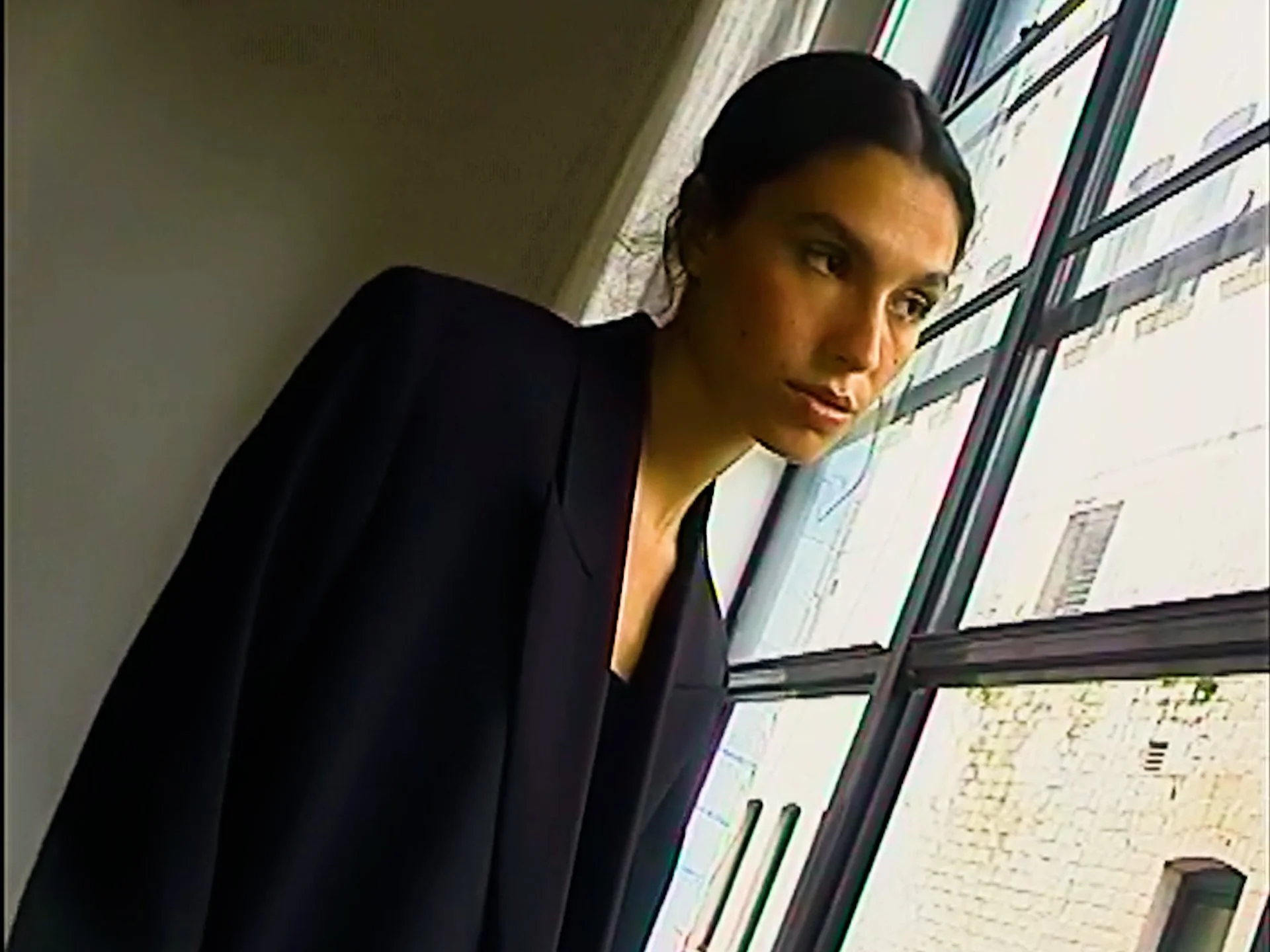 A woman with dark hair tied back, wearing a dark blazer, looking out a window with horizontal bars. Shot on a DV Camera kit through a digital recorder