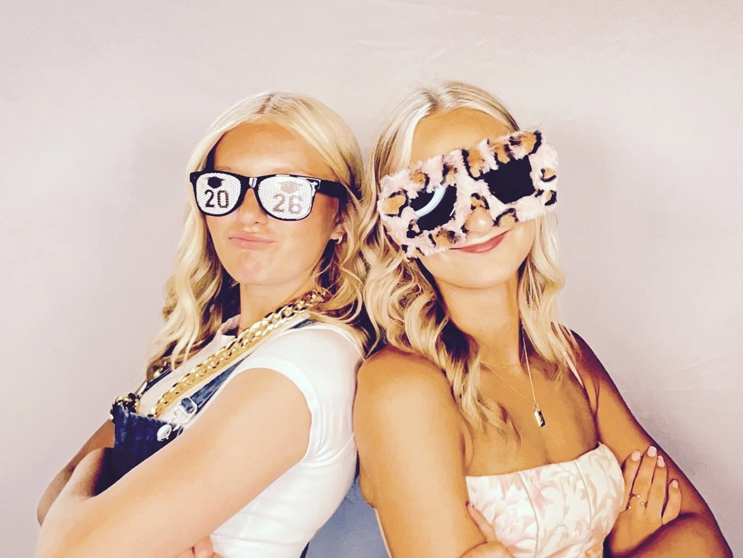 Two women with blonde hair wearing fashionable sunglasses, posing back-to-back with arms crossed, dressed in stylish outfits, celebrating a special occasion, possibly New Year's or a birthday.