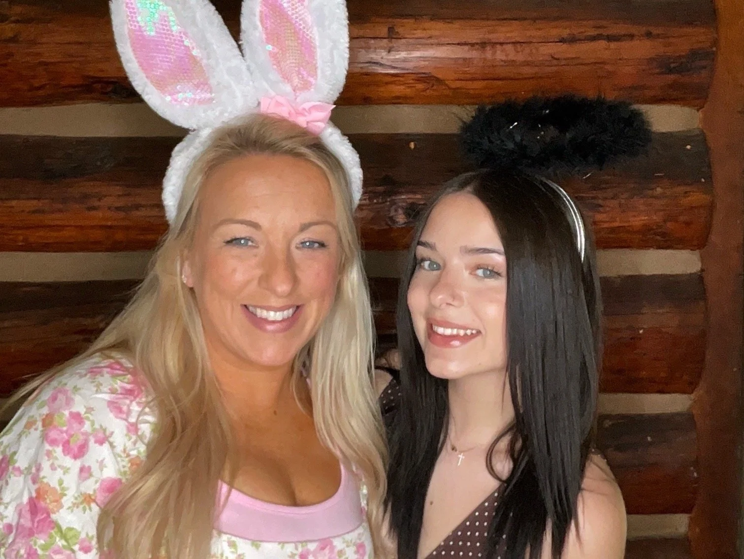 Two women dressed in Easter-themed costumes with bunny ears, smiling and posing for a selfie in front of a wooden wall.