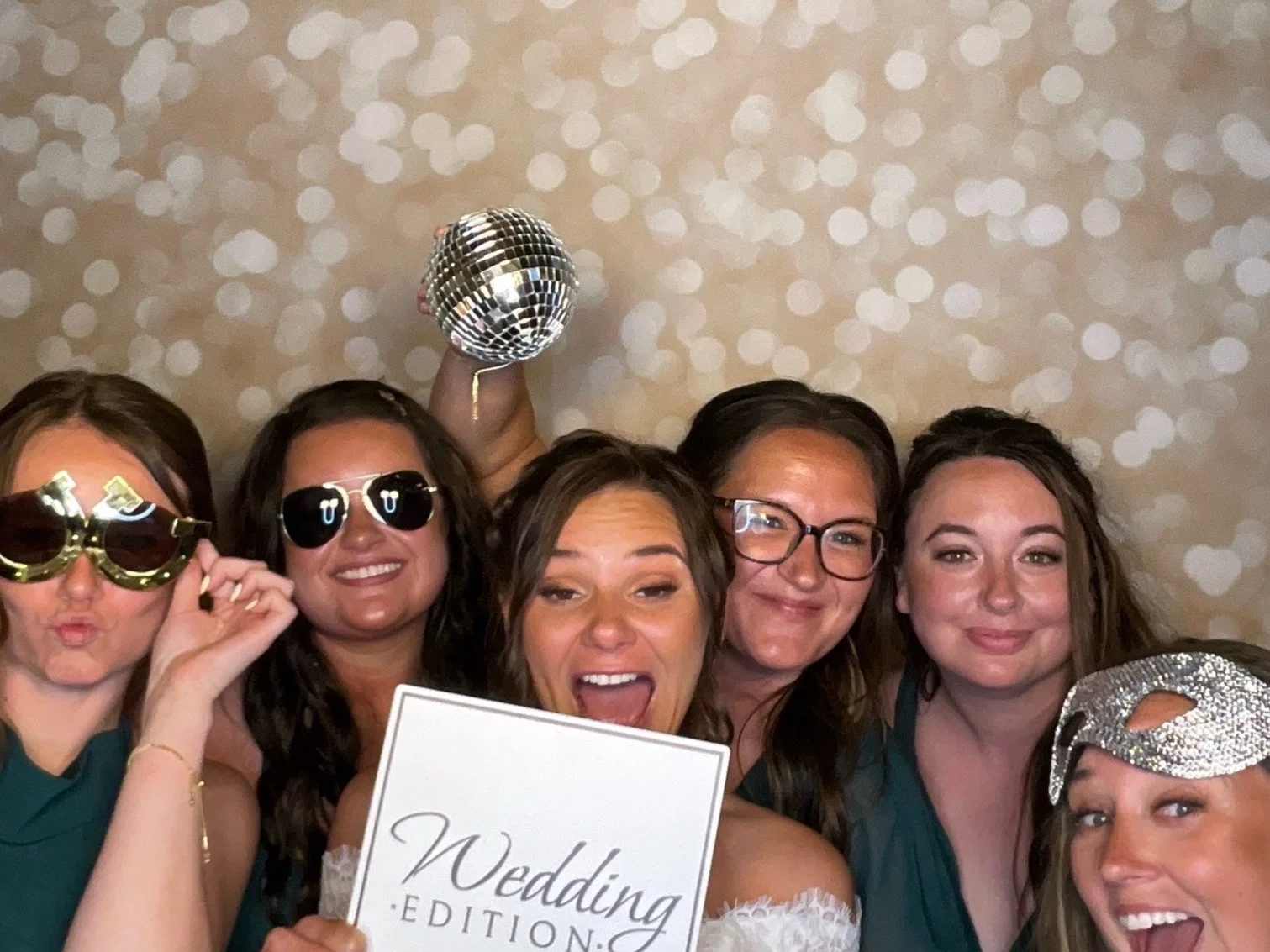 Group of five women celebrating at a wedding reception, wearing party glasses and holding a disco ball, with a sign that says 'Wedding Edition' in front of them, in front of a backdrop with a bokeh pattern.