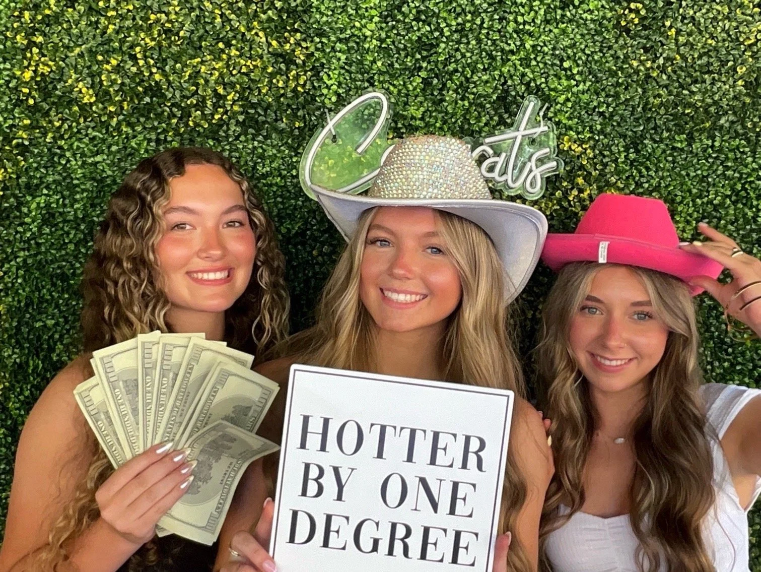 Three young women smiling at a celebration, standing in front of a green leafy backdrop. The woman on the left holds a fan of $100 bills. The woman in the middle wears a large white hat decorated with rhinestones and a neon sign that says 'Catt's.' The woman on the right wears a pink hat and is adjusting it. The woman in the middle holds a sign that reads 'HOTTER BY ONE DEGREE.'