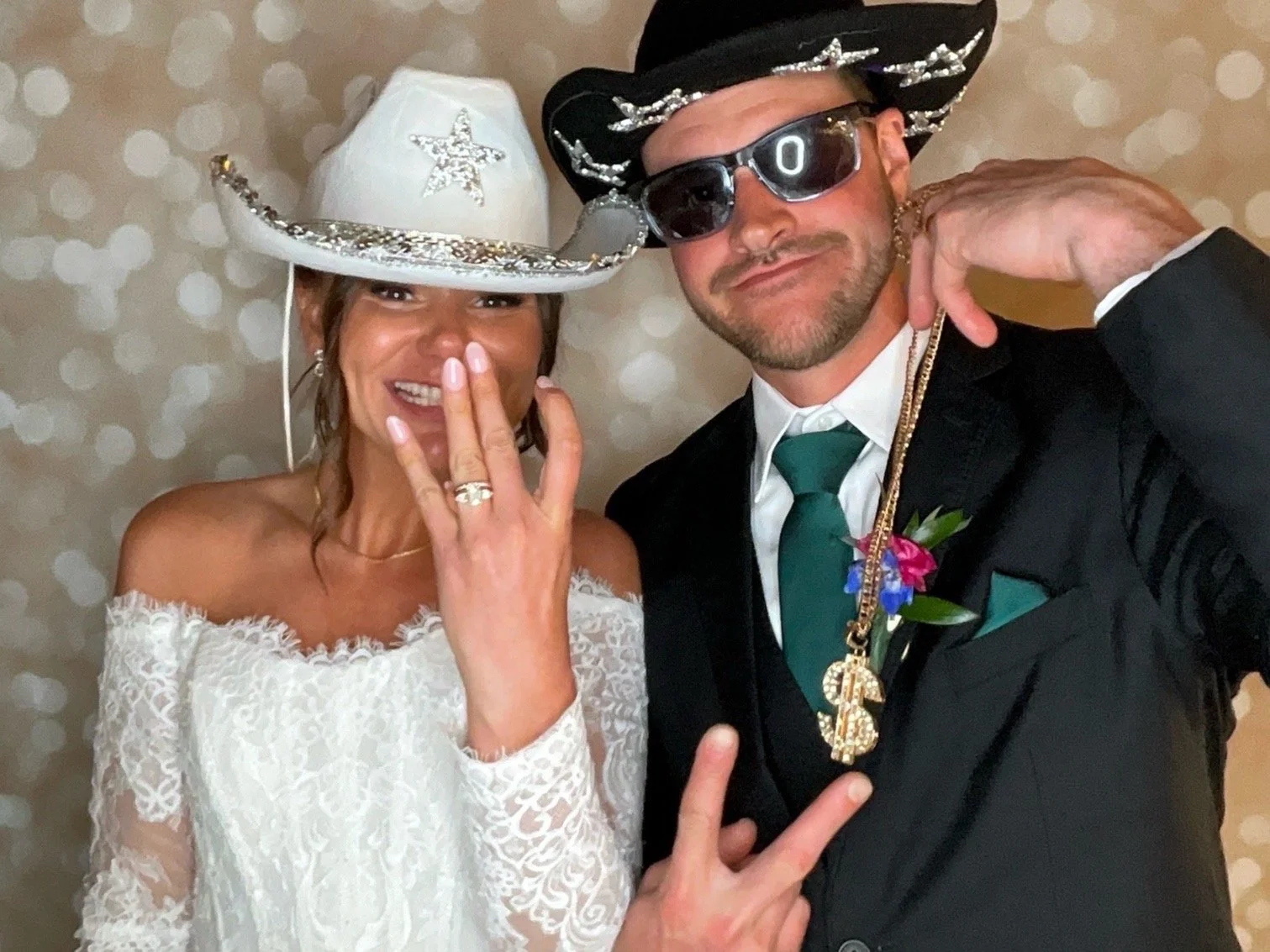 A woman and a man are dressed in cowboy-themed wedding attire. The woman is wearing a white lace off-shoulder wedding dress and a large white cowboy hat with silver star decorations, and she is showing her wedding ring. The man is wearing a black suit, white shirt, teal tie, and a black cowboy hat with silver stars, along with sunglasses and gold chains. They are posing in front of a beige polka dot background.