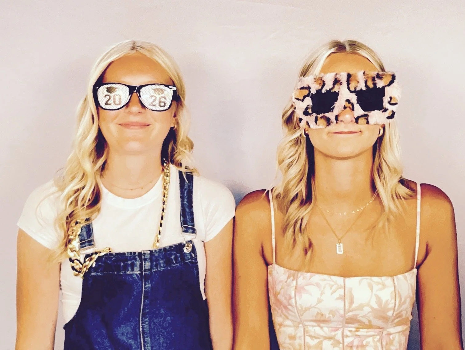 Two young women with blonde hair wearing fashionable outfits and fun glasses, standing against a plain background.