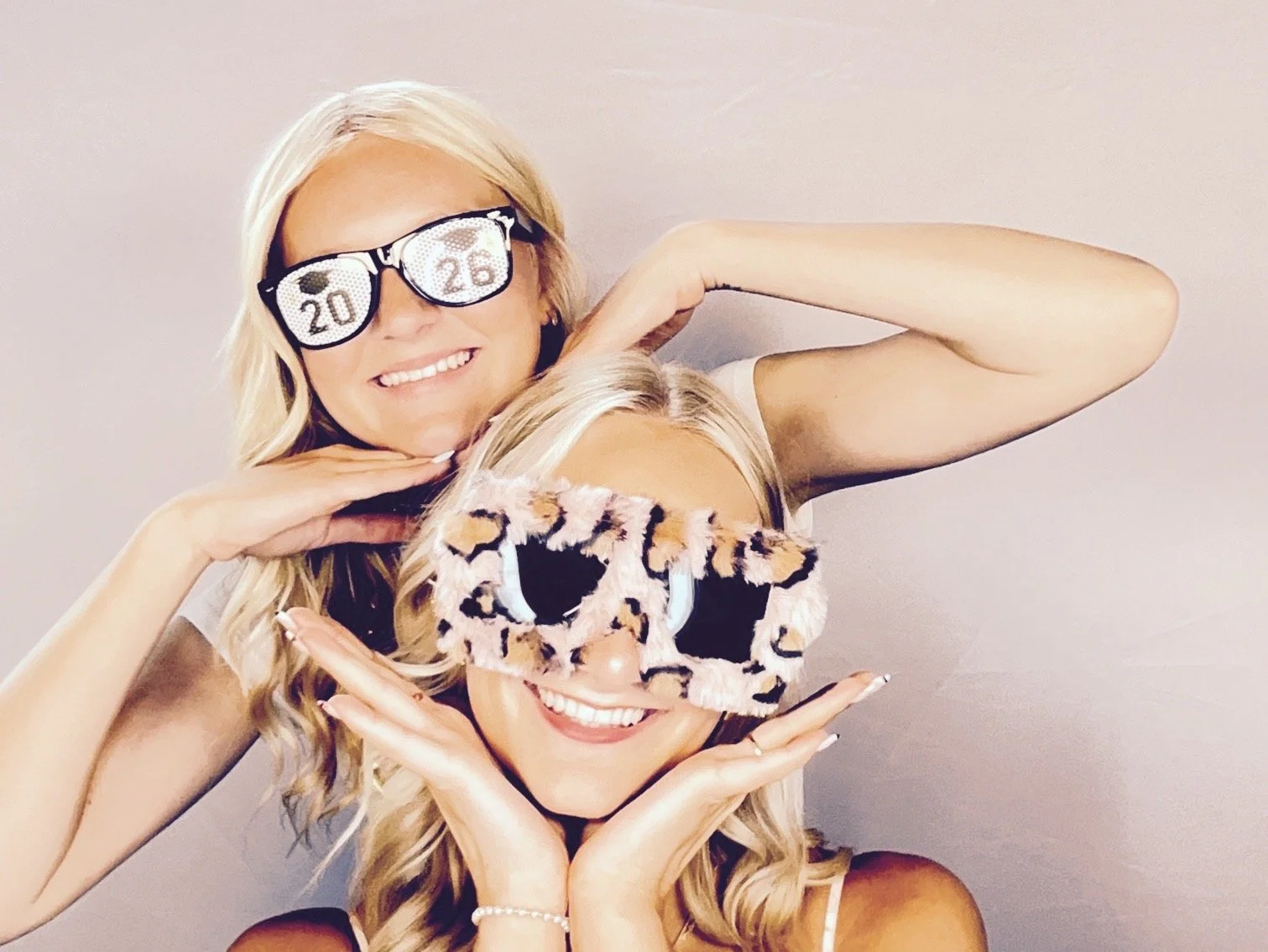 Two women with blonde hair wearing leopard print sunglasses, smiling and posing playfully with a beige background.
