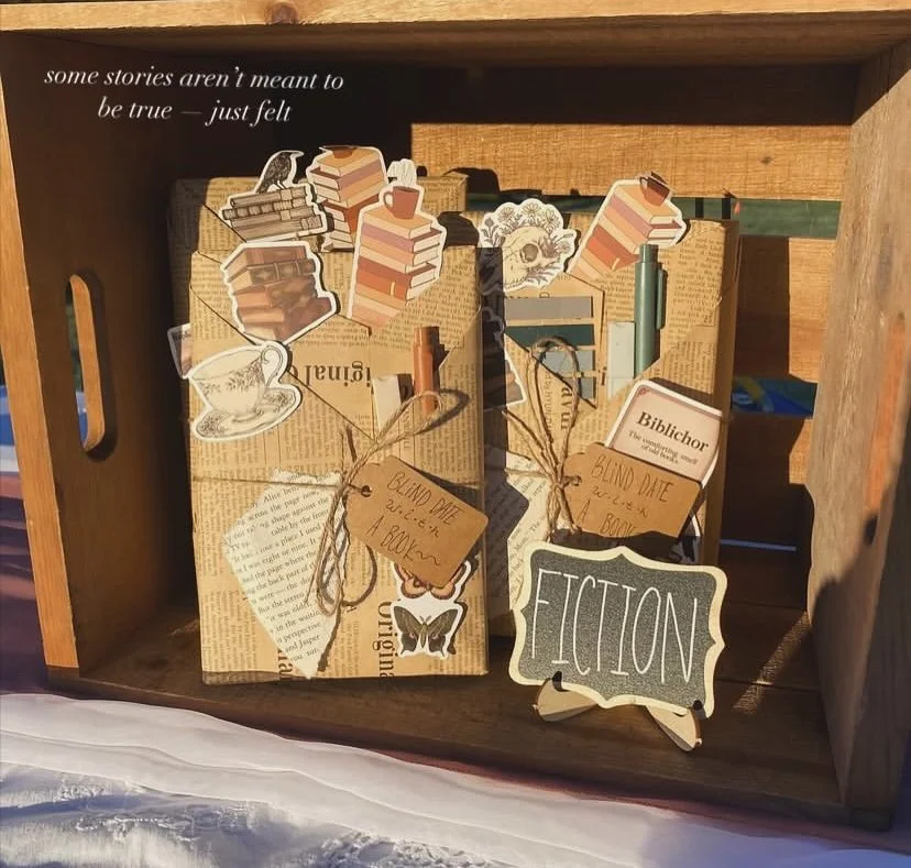 A wooden crate containing old books, stickers, and tags labeled "blind date with a book." There are illustrations of books, a teacup, a butterfly, and a chalkboard sign that says "Fiction."