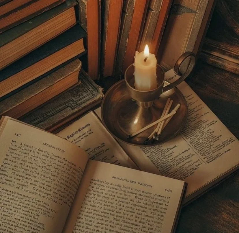 Open book on Shakespeare's writings, lit candle in metal holder, and sketchbook on wooden surface surrounded by books.