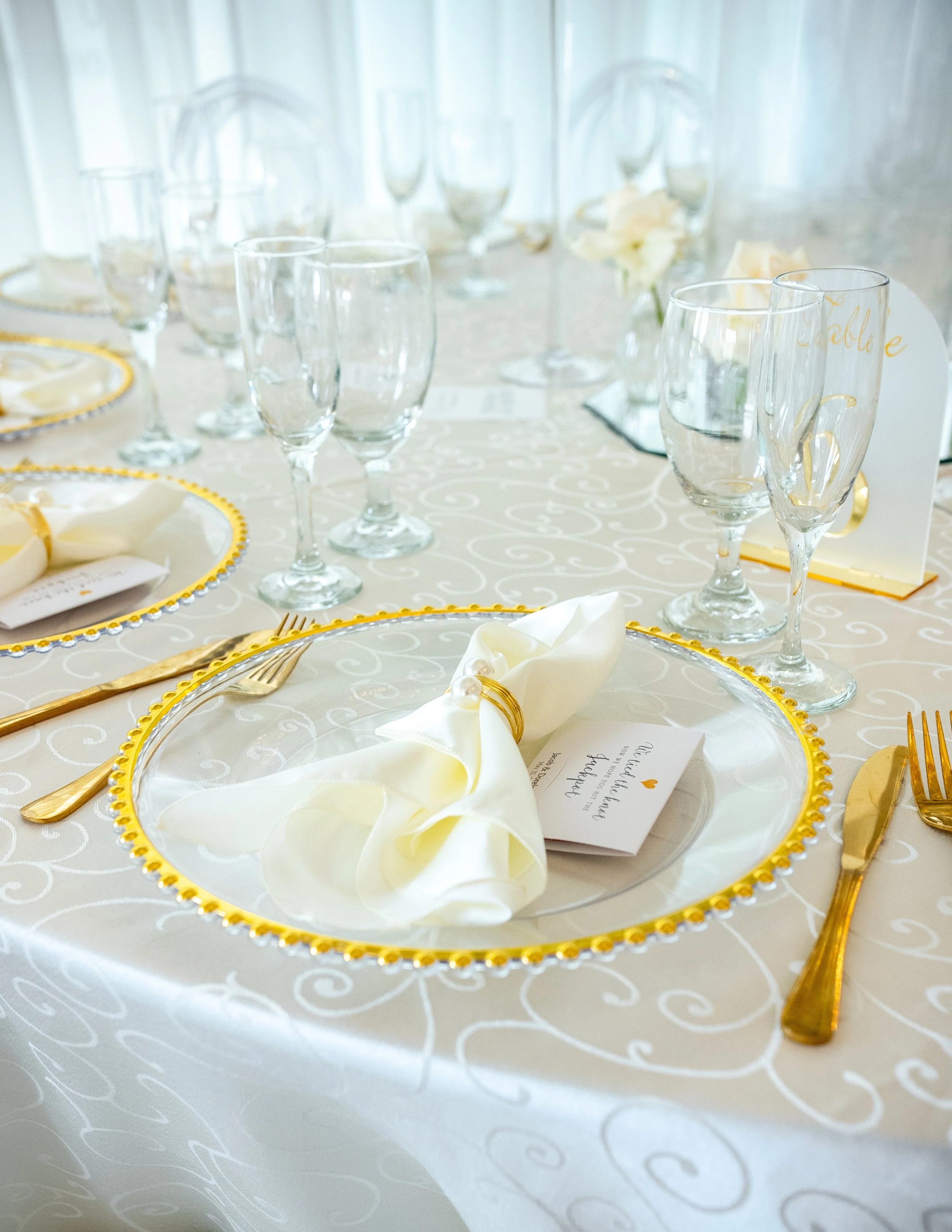 Elegant table setting with white and gold accents, multiple empty wine glasses, gold flatware, white napkin with a pearl ring, and a menu card.