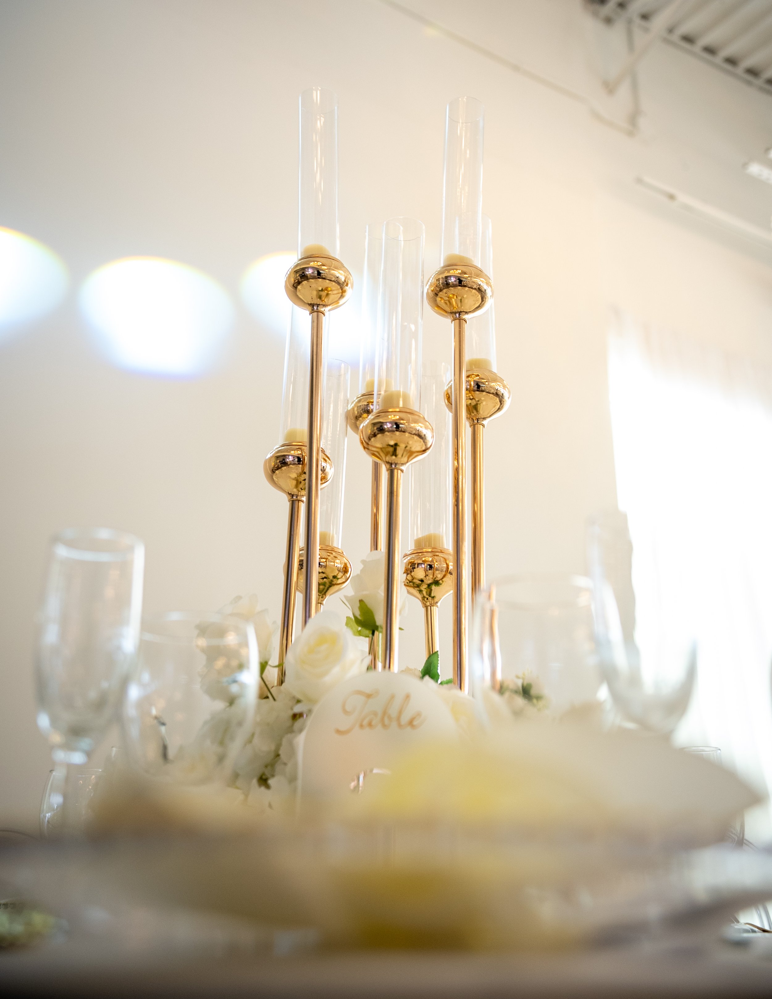 Elegant table centerpiece with tall glass cylinders and gold spheres, surrounded by white flowers and glassware at a wedding or special event.
