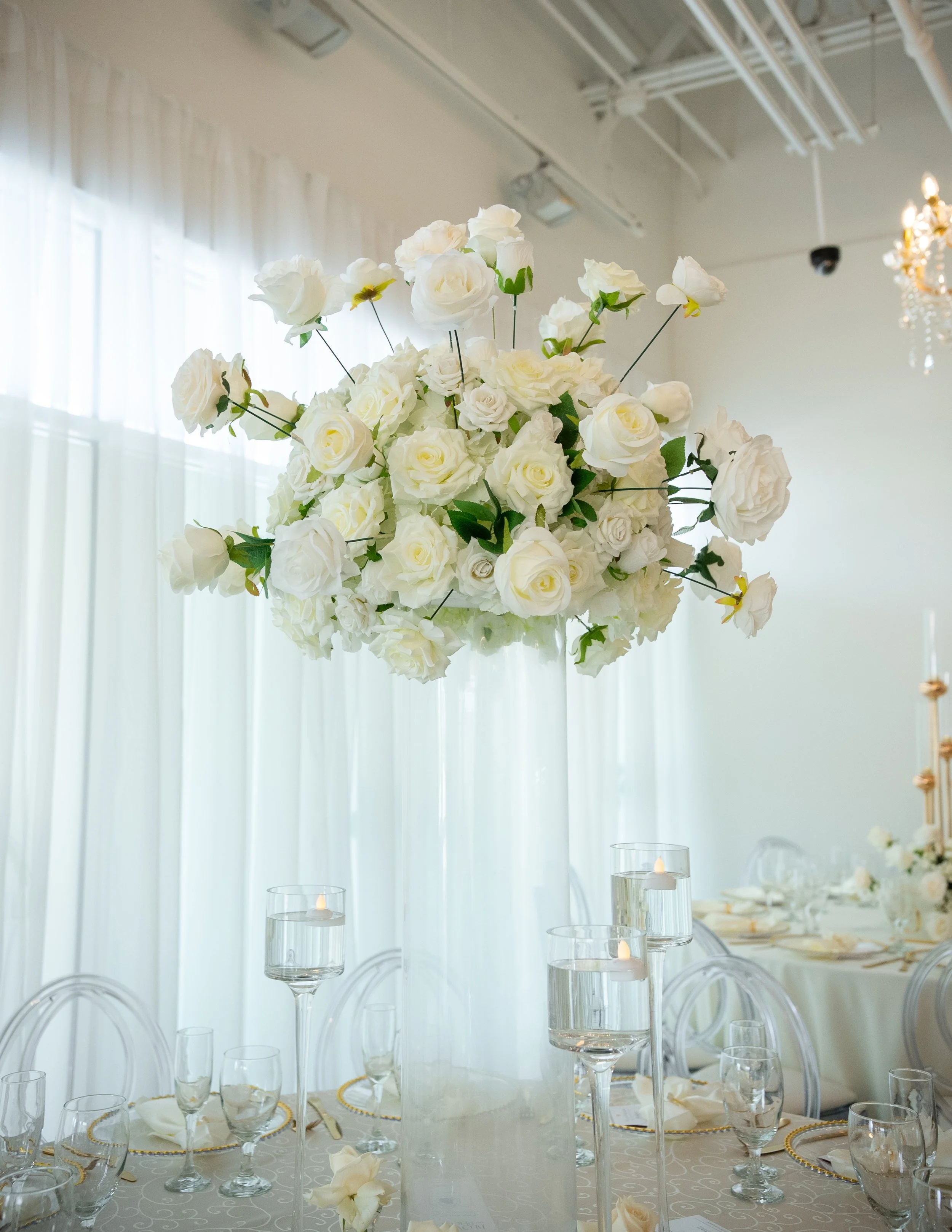 Elegant tall floral centerpiece with white roses and greenery on a dining table in a bright, decorated event space with white curtains and chandelier.