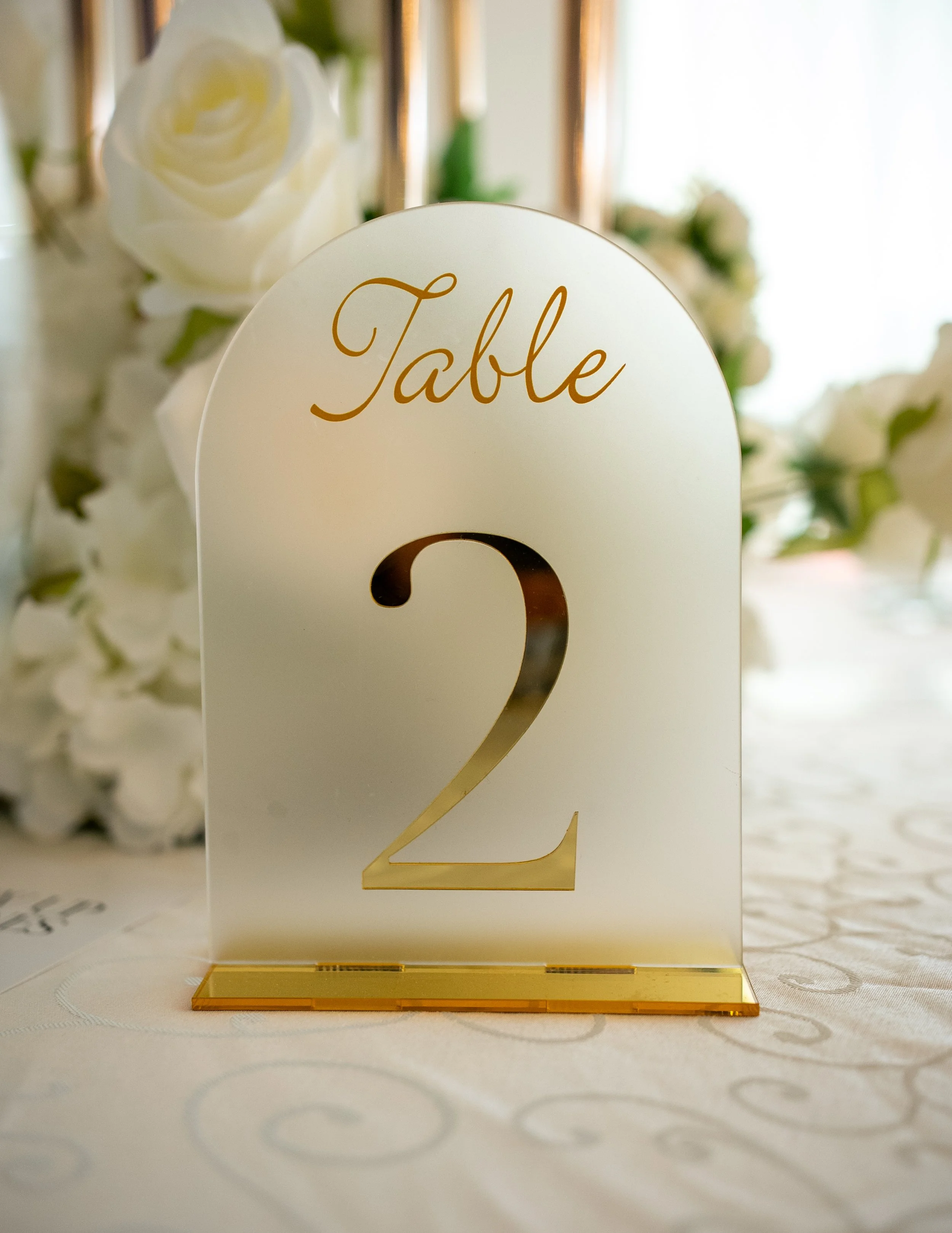 Table number sign with the number 2 on a gold base, wedding or event decor with white roses in the background.