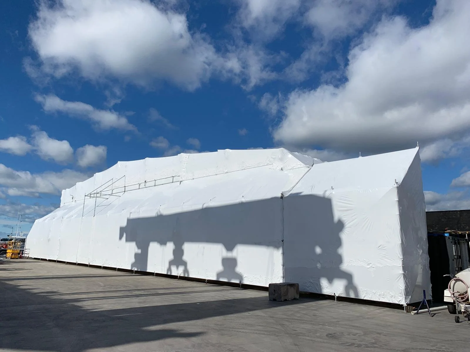 Large white tent or temporary structure with a shadow cast on it, set outdoors on a clear day with blue sky and scattered clouds.