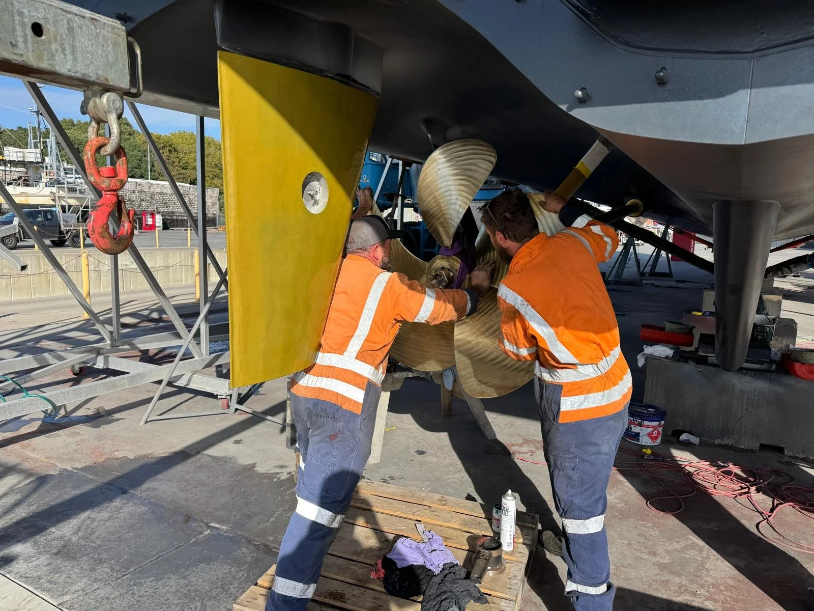 Two workers in orange safety uniforms working on a boat's propeller at a marina, with tools and equipment nearby.