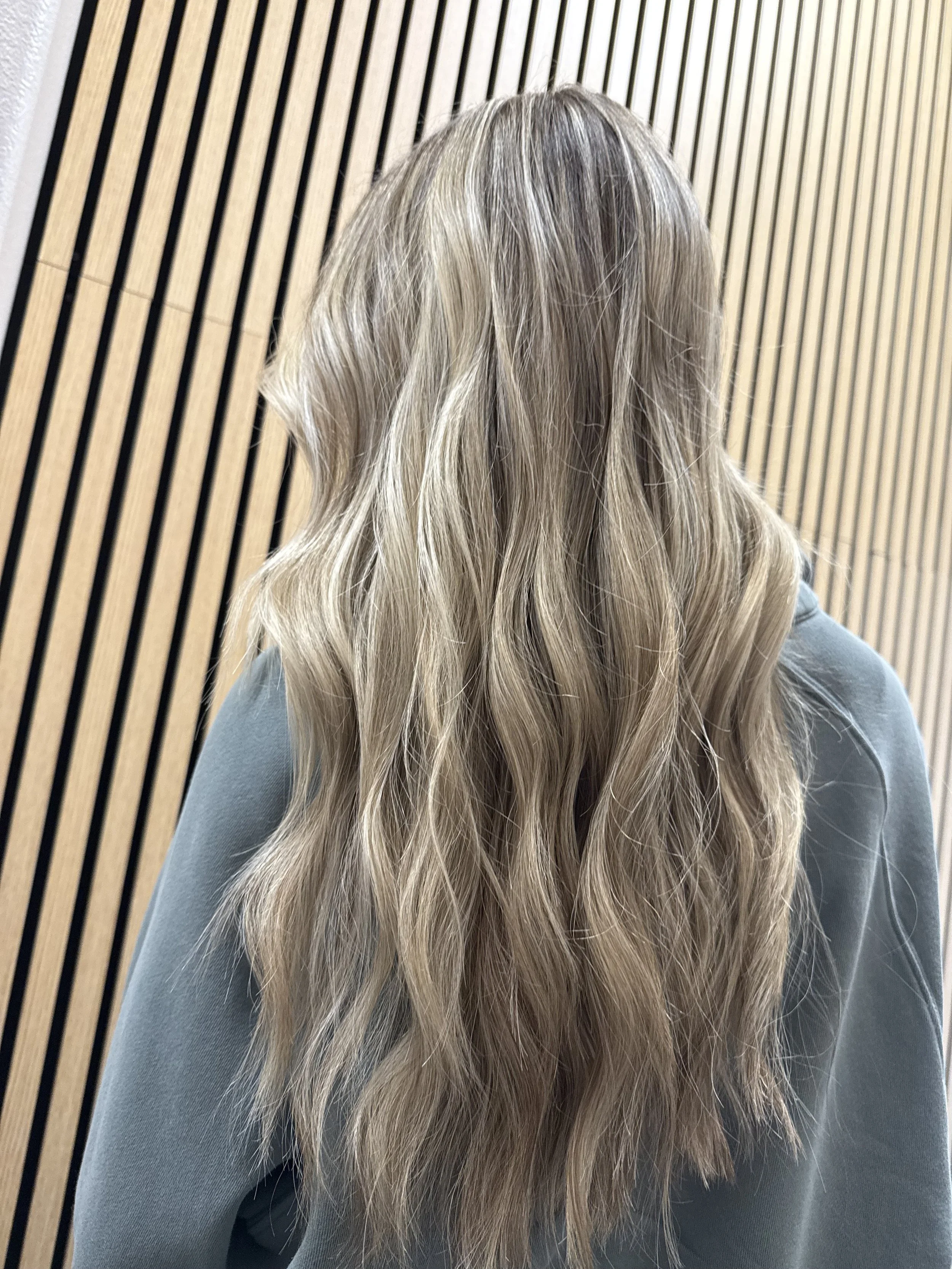 Back view of a woman with long, wavy blonde hair standing against a wooden slatted wall.