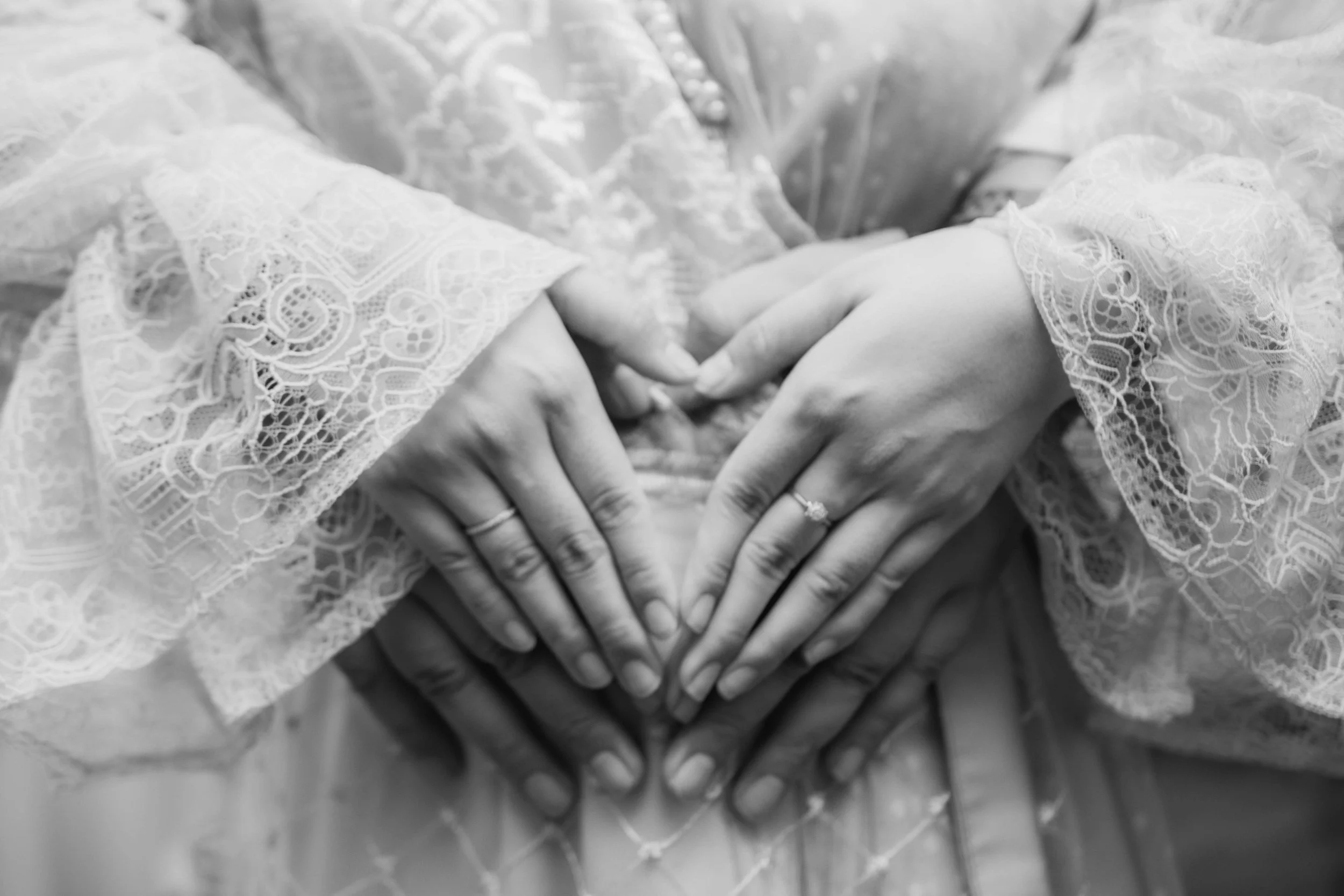 Close-up of two women forming a heart shape with their hands, wearing lace and engagement rings, in black and white.