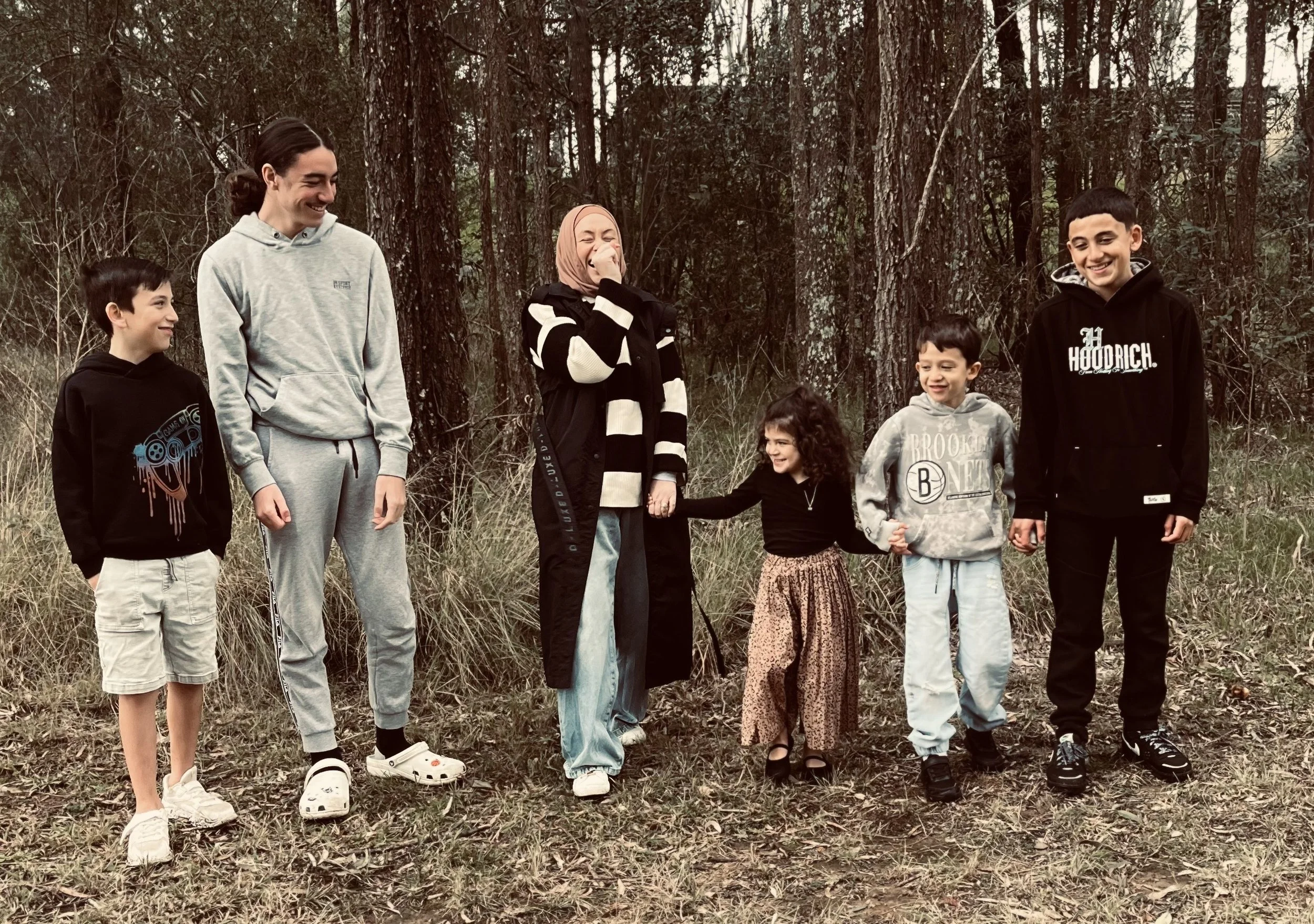 Seven people, including three children and four adults, standing in a line outdoors among trees and grass, holding hands and smiling, during a casual gathering.