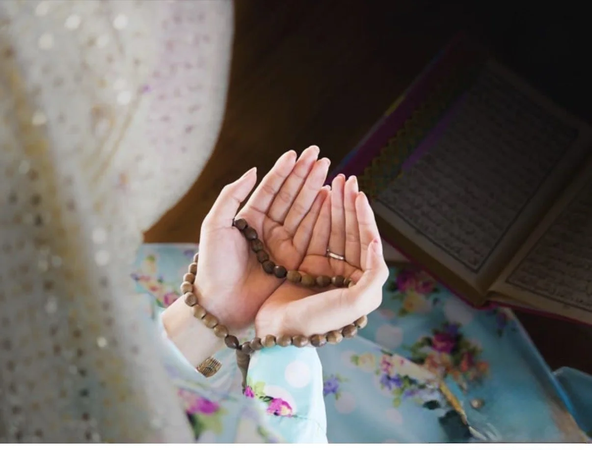 A person holds prayer beads and a prayer book open, sitting on a colorful cloth.