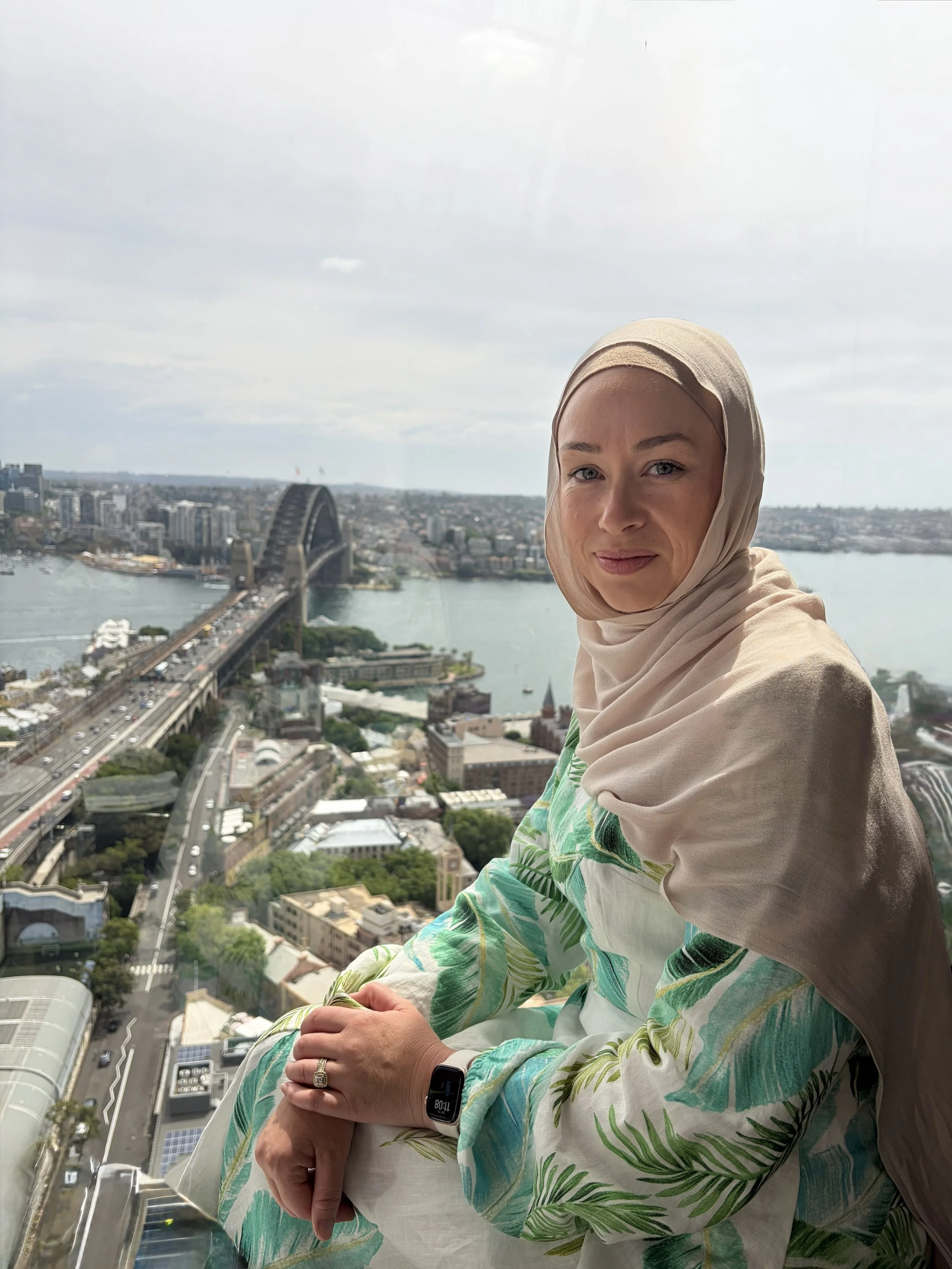 A woman wearing a beige hijab and a tropical print dress sits against a glass window, overlooking the Sydney Harbour Bridge and the city skyline in the background.