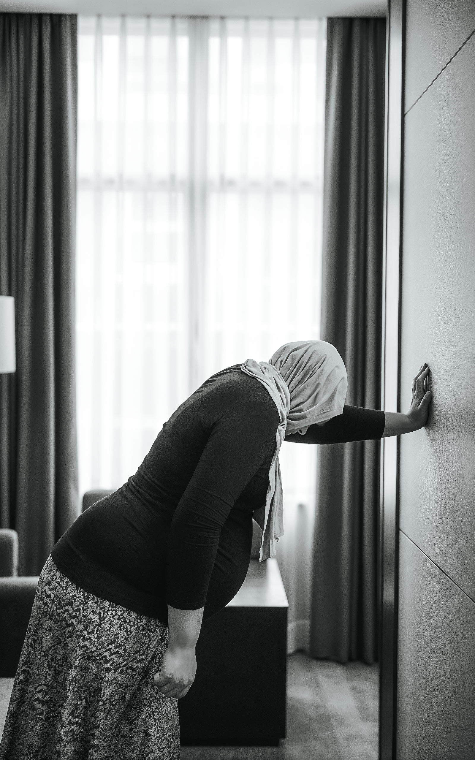 A woman wearing a hijab is leaning against a wall in a hotel room, with her head bowed and one hand touching the wall.