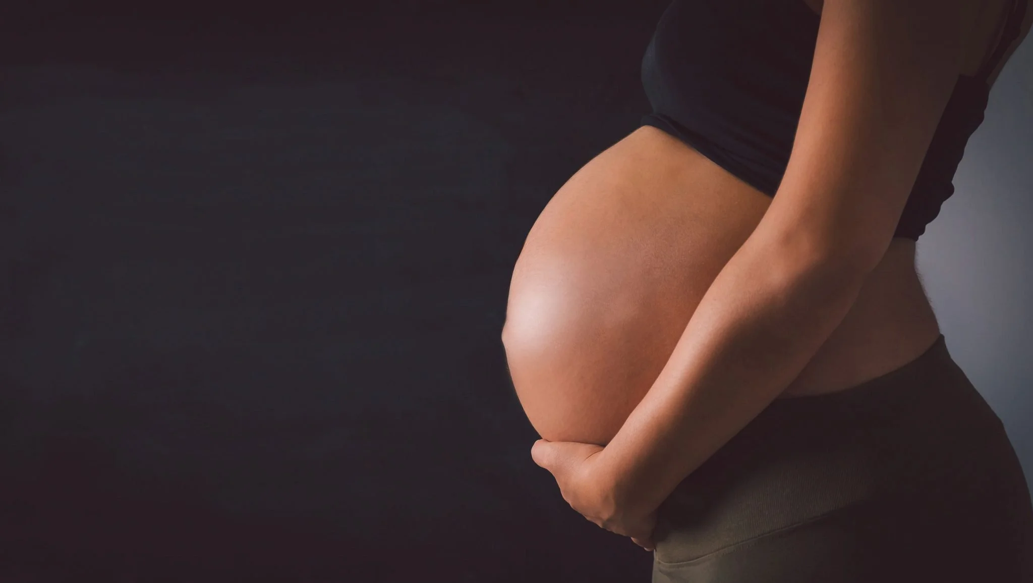 A pregnant woman lightly holding her rounded belly with both hands, wearing a black top and dark pants, against a plain, dark background.