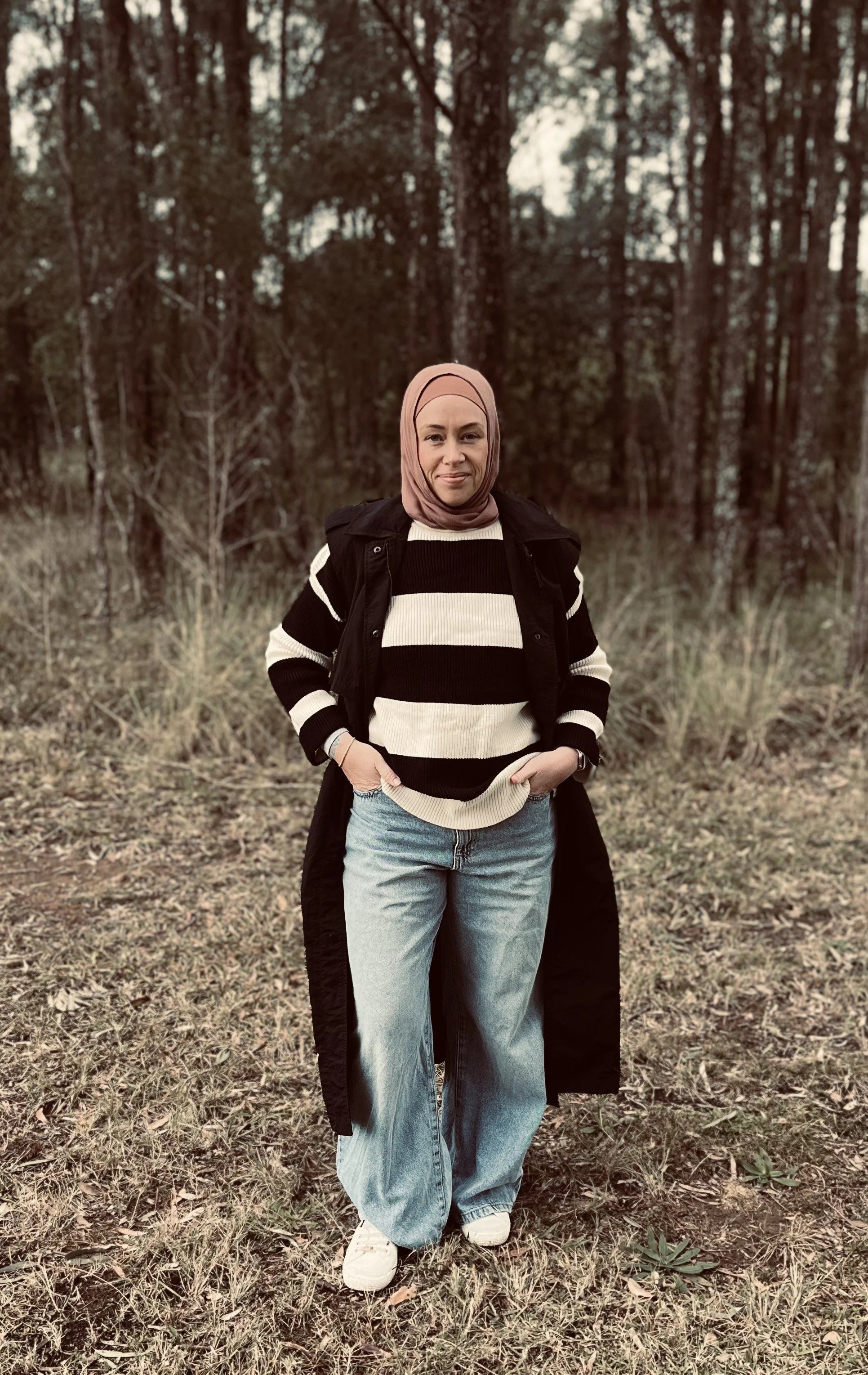 A woman wearing a beige hijab, black and white striped sweater, black jacket, light blue jeans, and white sneakers stands outdoors on grass in front of a wooded area.