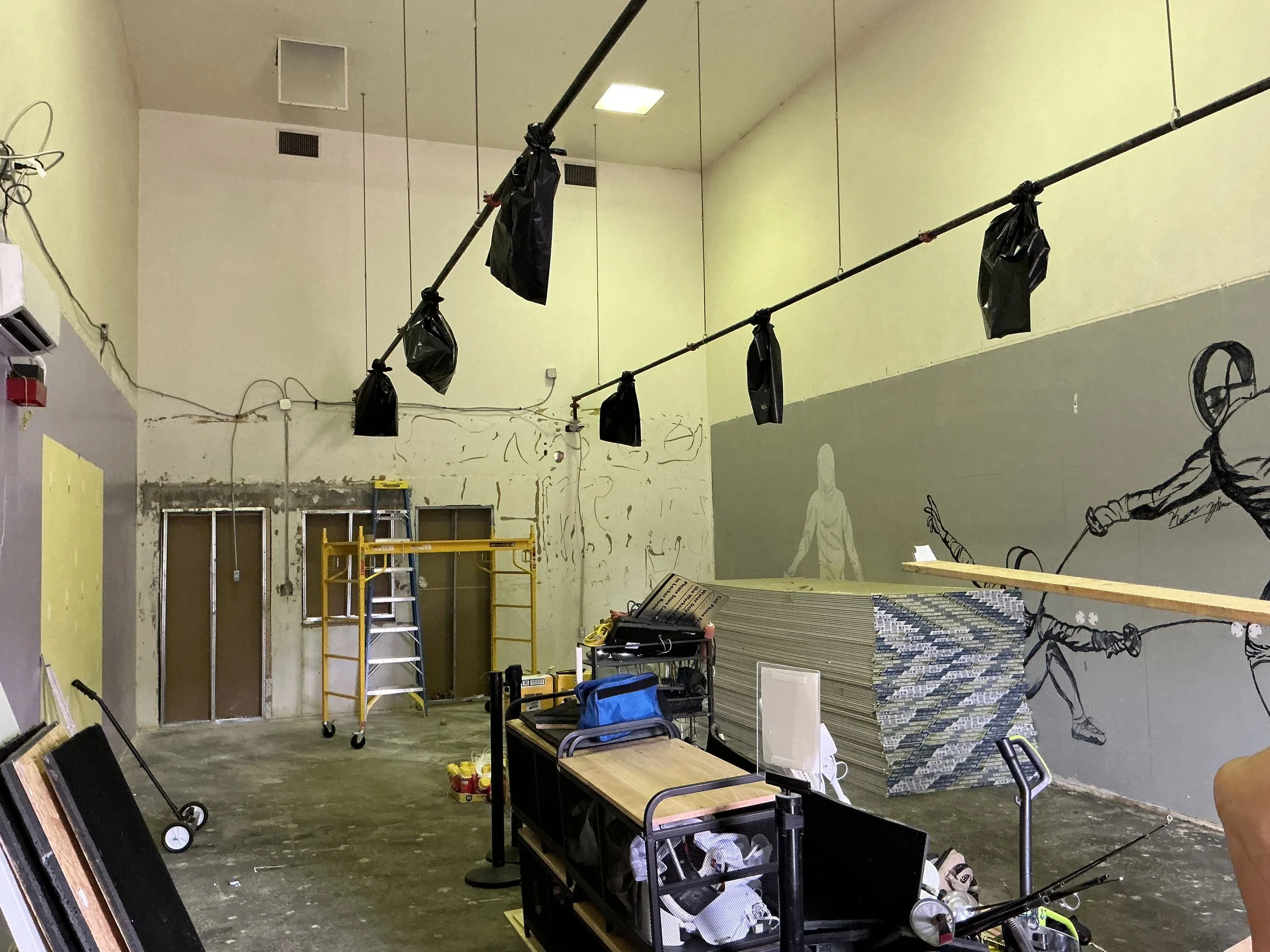 An interior room under construction with various tools, equipment, and shelving. Black bags hang from a metal rod across the room. The wall has sketches and incomplete murals, including a sketch of a person and a figure with an outstretched arm.